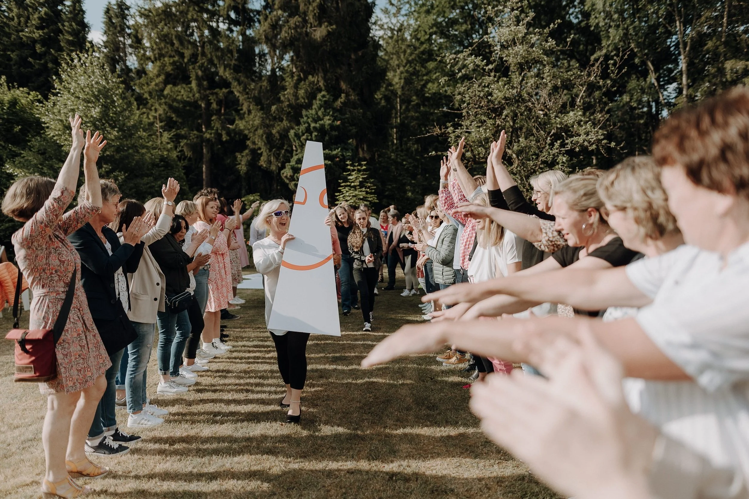 Eine Gruppe von Menschen steht im Freien in zwei Reihen und bildet eine Art Spalier. Eine Person in der Mitte trägt ein großes, lächelndes Gesichtsschild. Die Gruppe scheint zu feiern oder zu applaudieren. Im Hintergrund sind Bäume und ein blauer Himmel zu sehen.