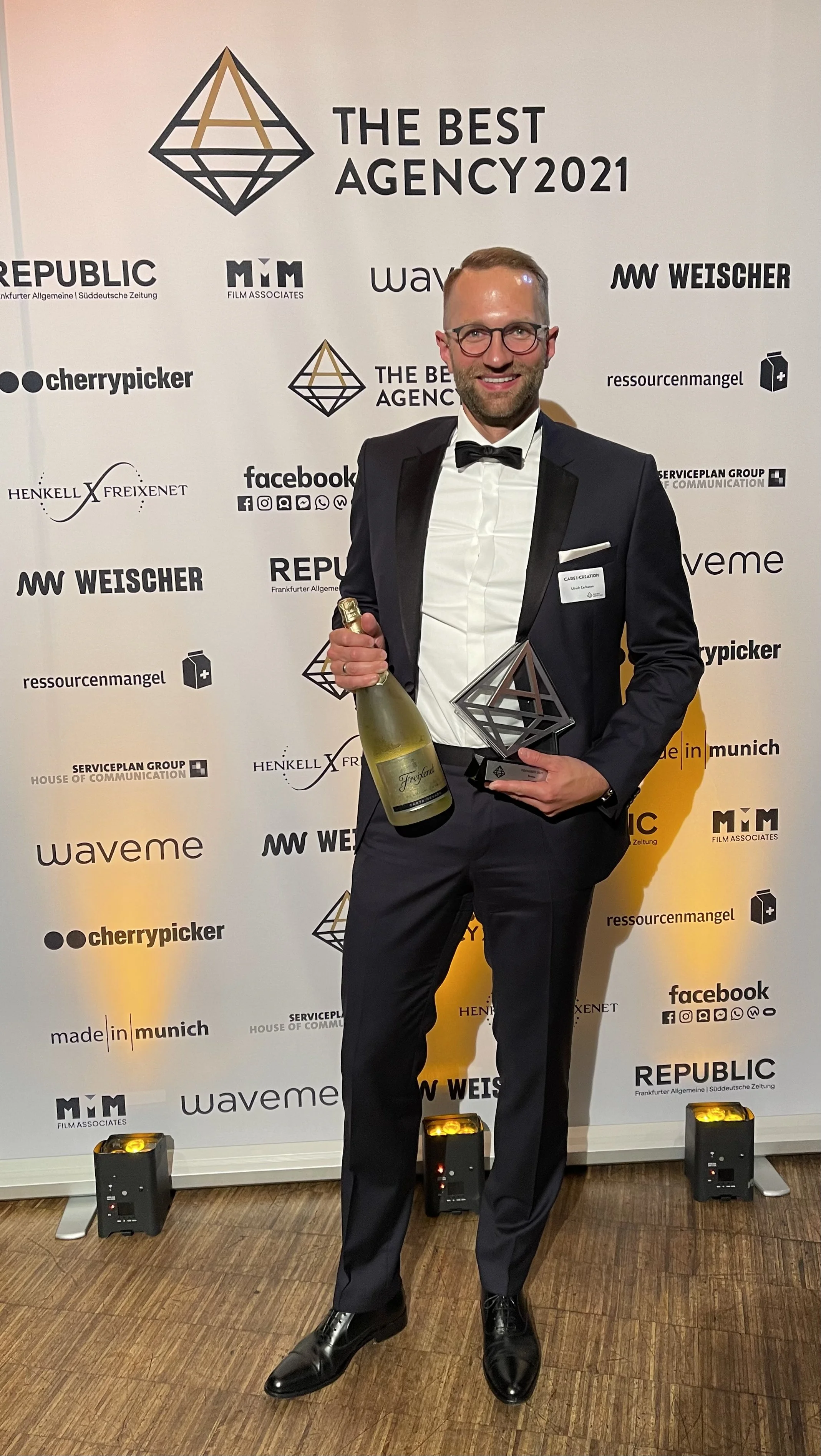 Ulrich Zerhusen hält eine Flasche Sekt in der Hand und die Trophäe bei einer Preisverleihung des The Best Agency Award, Hintergrund mit Logos und Texten.