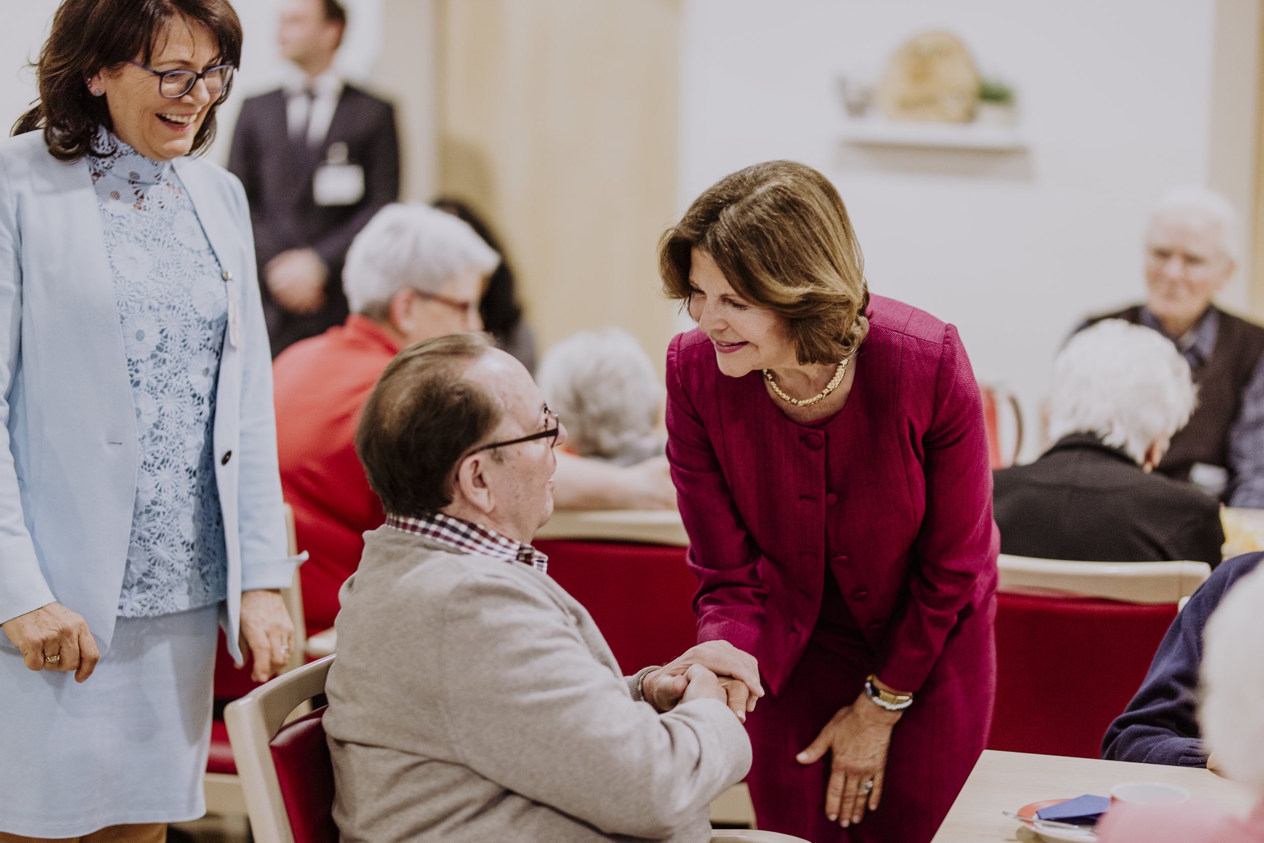 Eine Frau in einem roten Kleid schüttelt die Hand eines sitzenden älteren Mannes, während eine andere Frau in einem hellblauen Outfit zusieht. Die Szene findet in einem Raum mit mehreren Menschen statt.