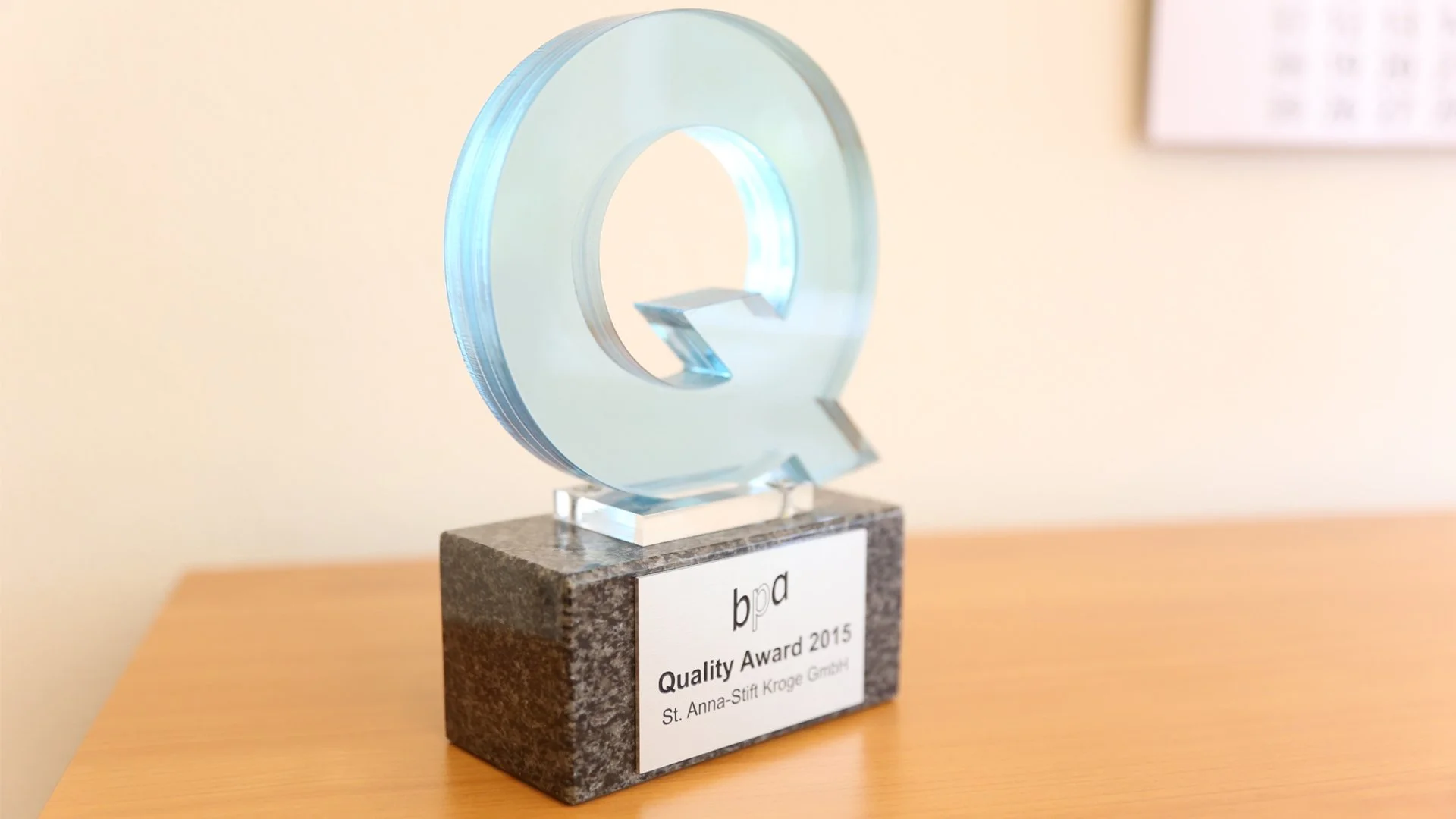 Glas-Preisbrosche für den Qualitätsaward 2015, von st. anna-stift kroge gmbh, auf grauem Sockel auf Holztisch vor hellem Wand