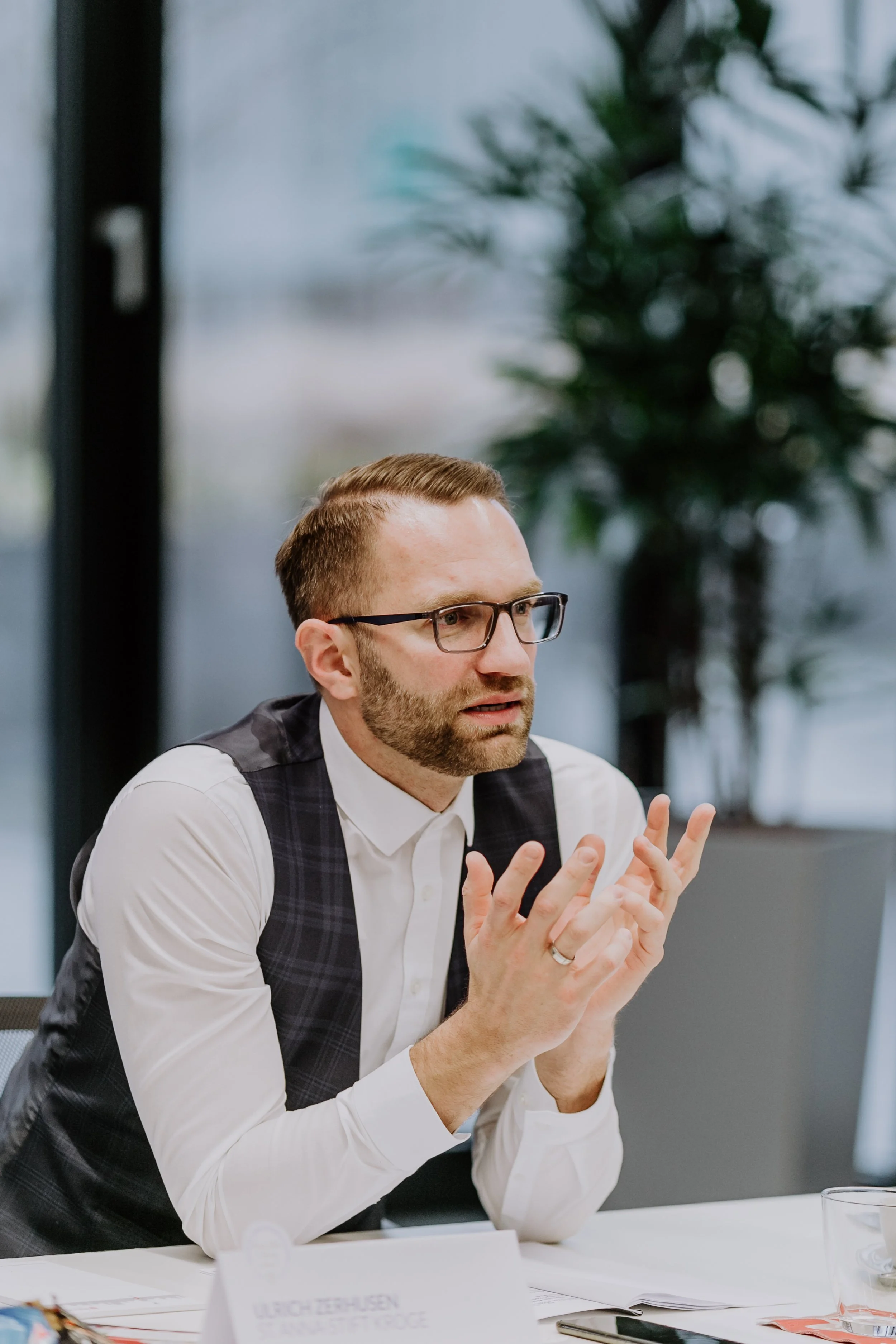 Ein Mann mit Brille, weißen Hemd und Weste sitzt an einem Tisch in einem Besprechungsraum, er sieht nachdenklich aus, während er mit den Händen gestikuliert und spricht.