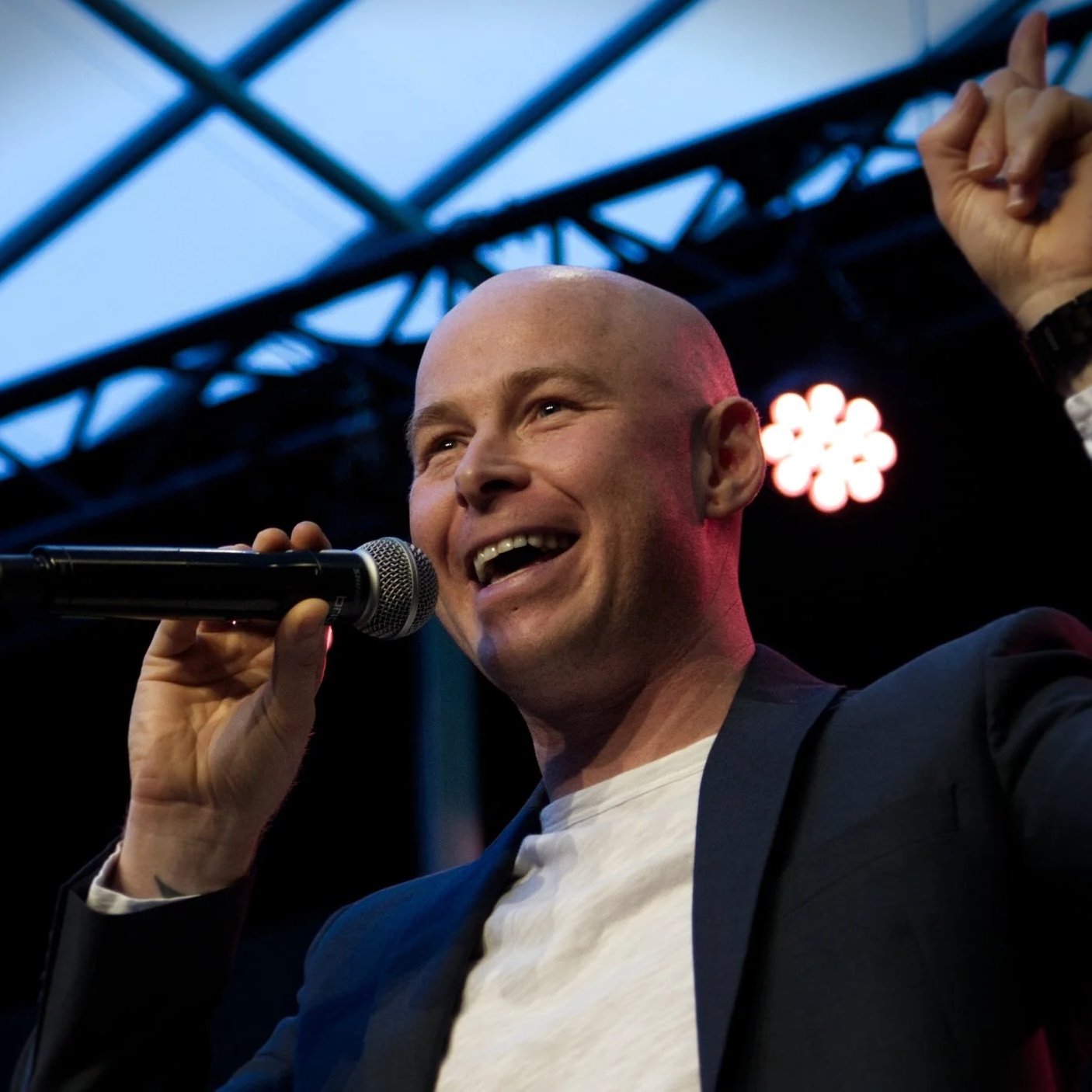 A man with a shaved head speaking or singing into a microphone on stage, raised hand making a fist gesture, stage lighting and scaffolding visible in the background.