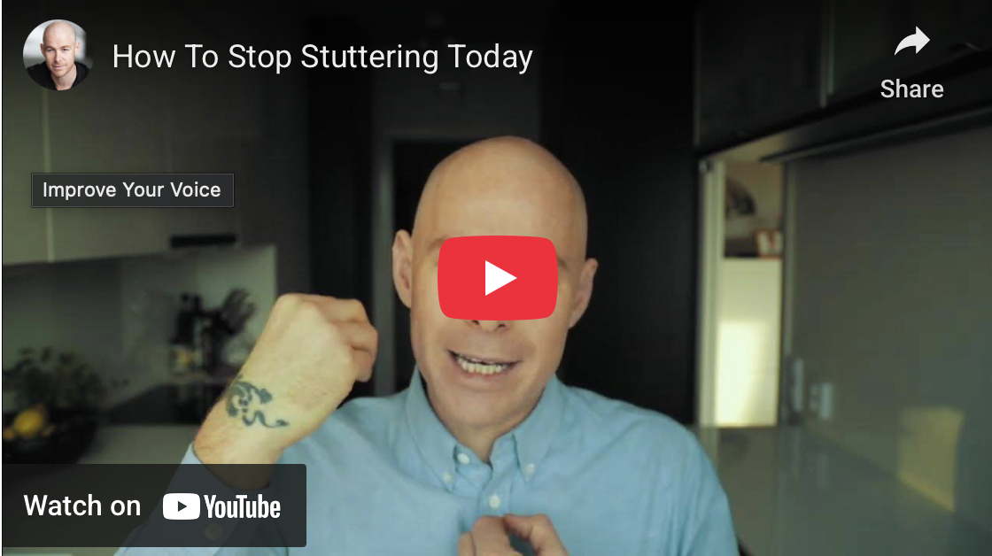 A man with a shaved head wearing a light blue button-up shirt, gesturing with his fist near his face, in a kitchen with cabinets and a TV.