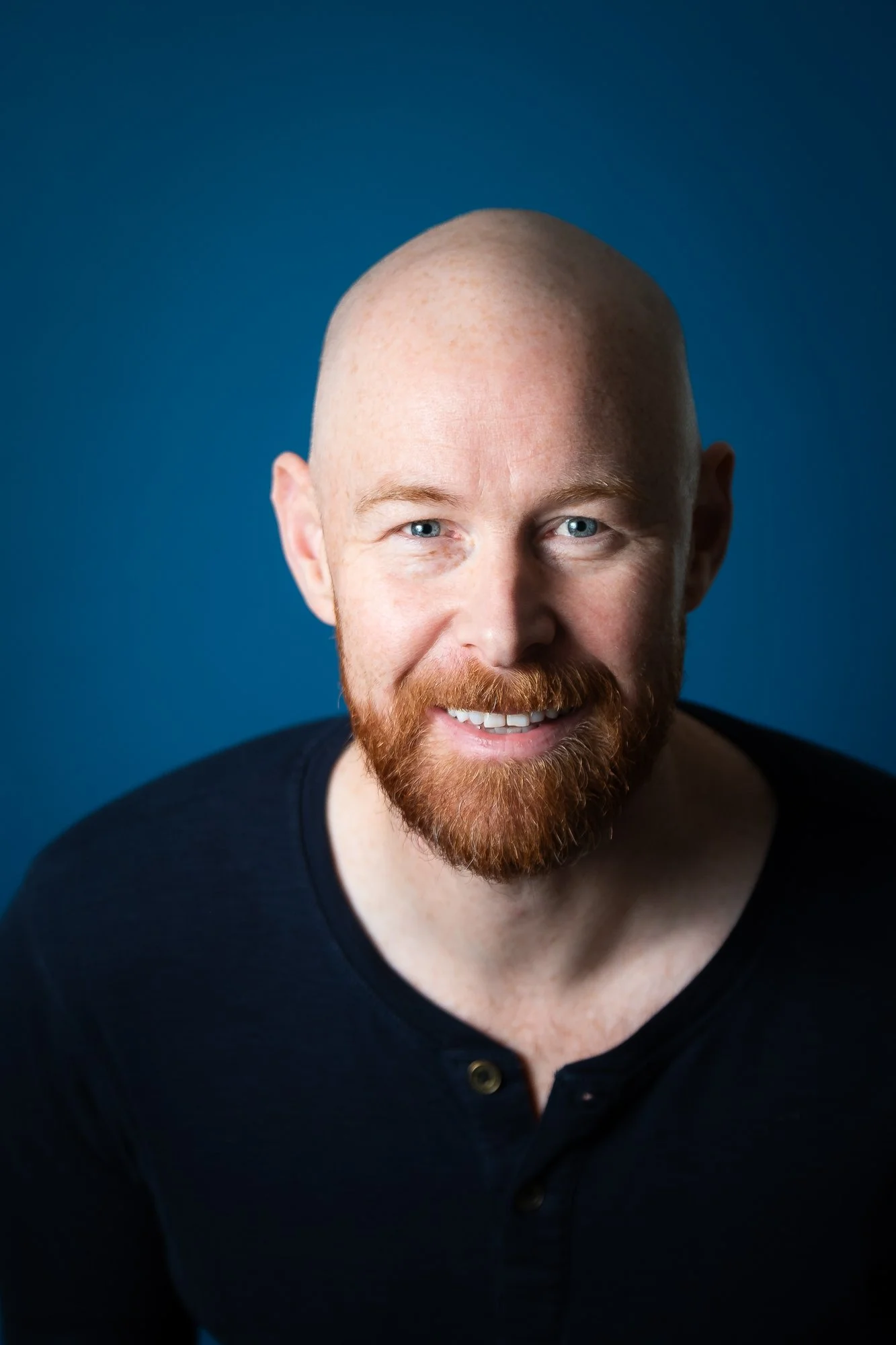 Headshot of a smiling man with a red beard and blue eyes, wearing a black shirt, against a blue background.