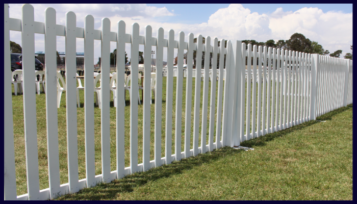 Long stretch of white picket fencing set up on a grassy field with white chairs in the background, commonly used for events or temporary boundaries.