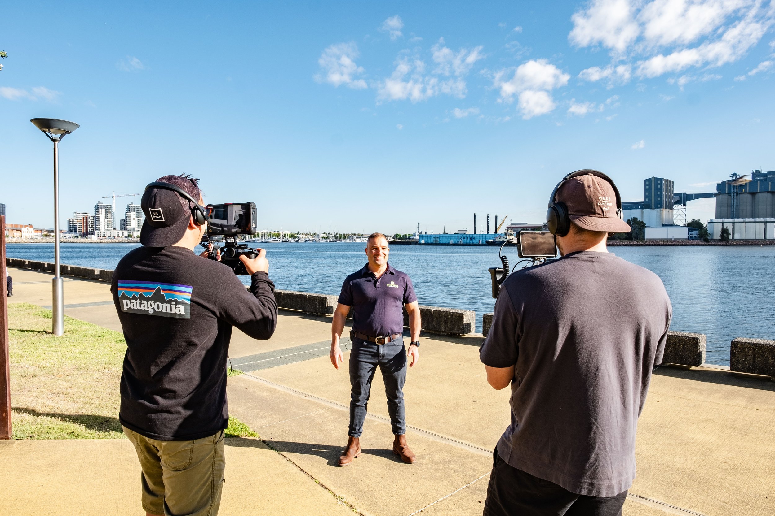 Two cameramen filming a person by a waterfront with industrial buildings in the background on a sunny day.