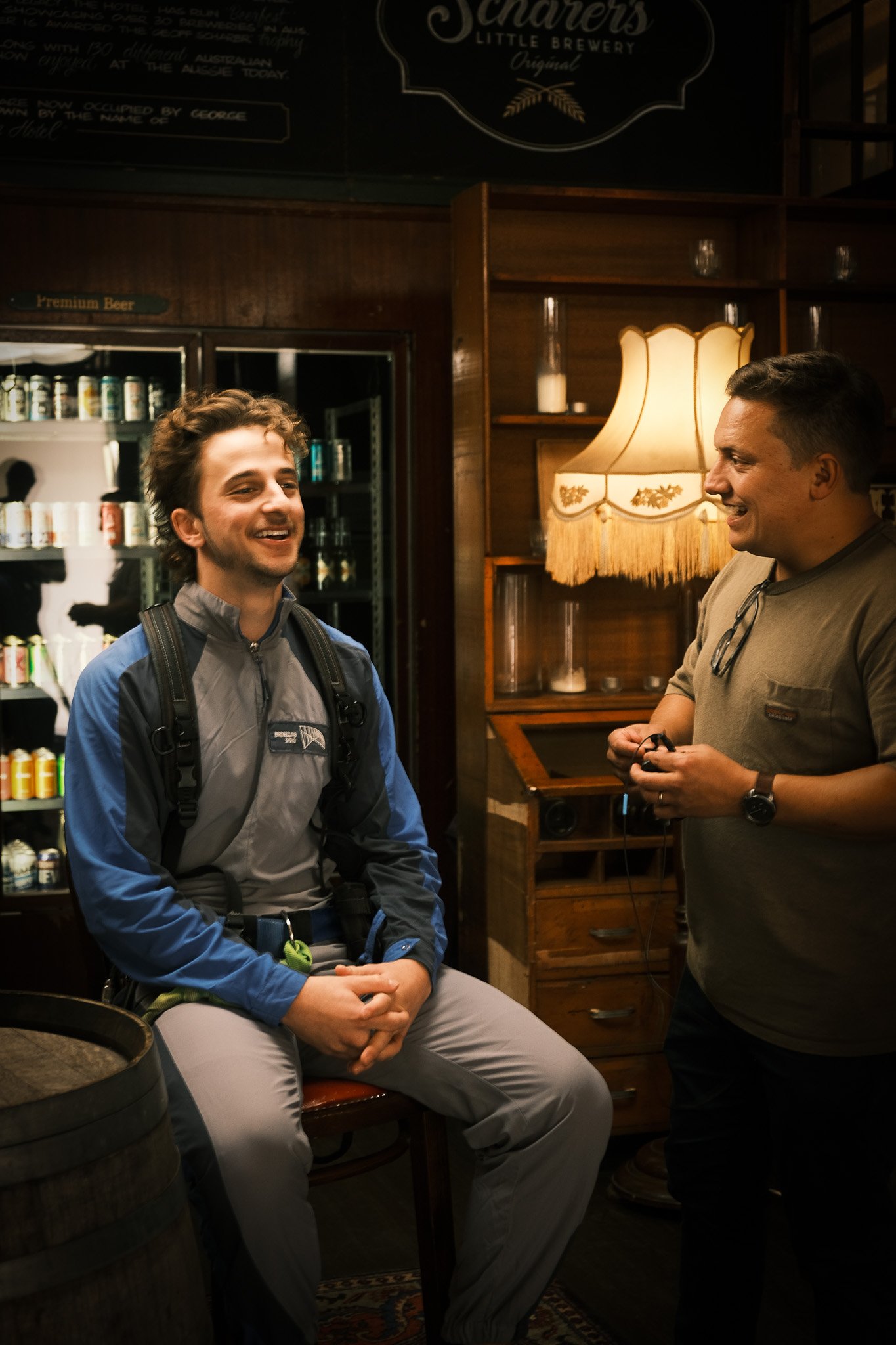 Two men in a cozy pub setting, one seated wearing casual outdoor gear, and the other standing with a smile, engaged in conversation. A vintage lamp and shelves with bottles and candles are in the background.