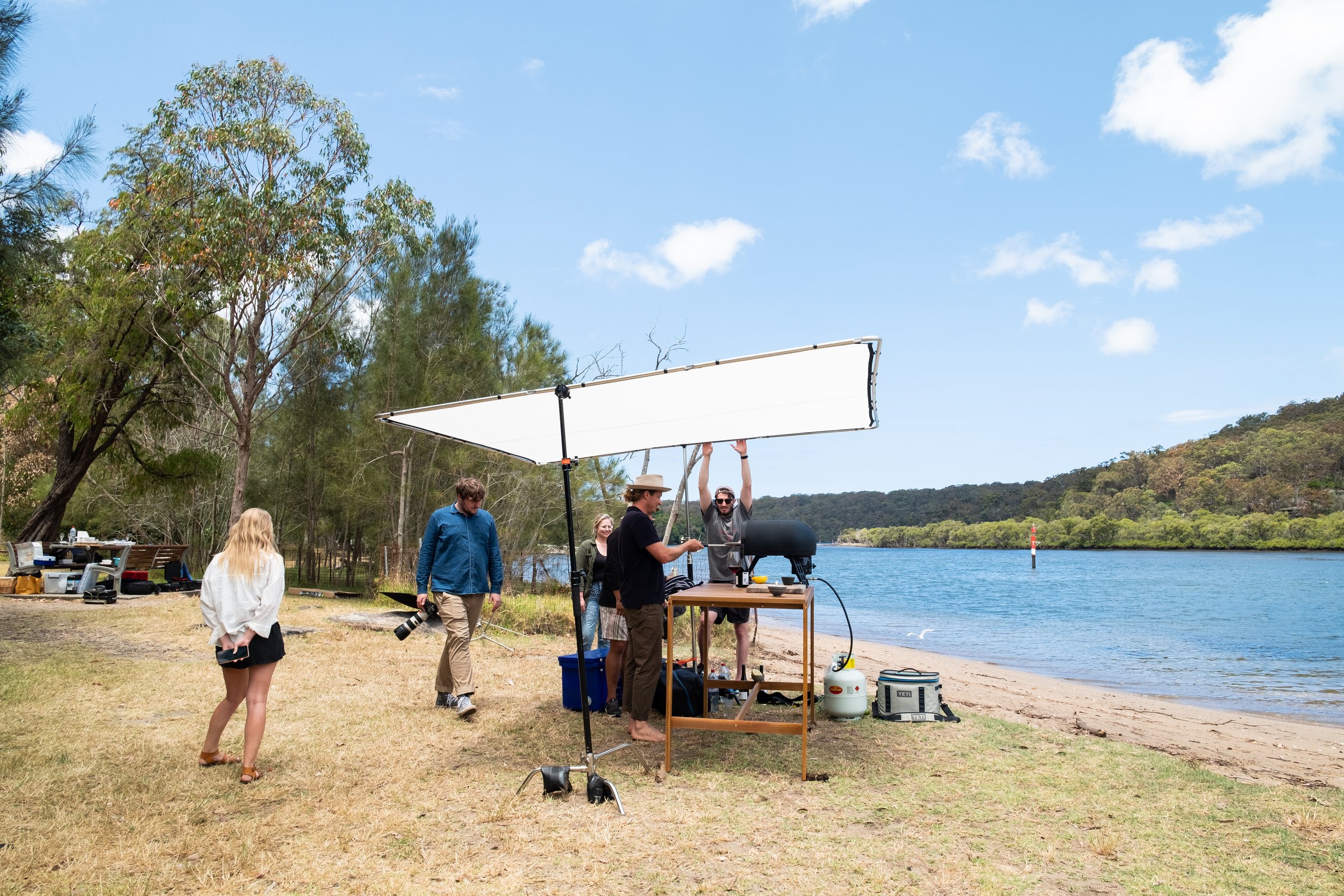 People setting up a filming scene by a river, with lighting equipment and a canopy, surrounded by trees and grass on a sunny day.