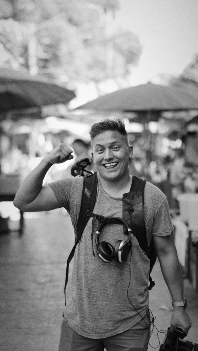 Man in gray shirt with headphones around neck, smiling and flexing arm in outdoor market.