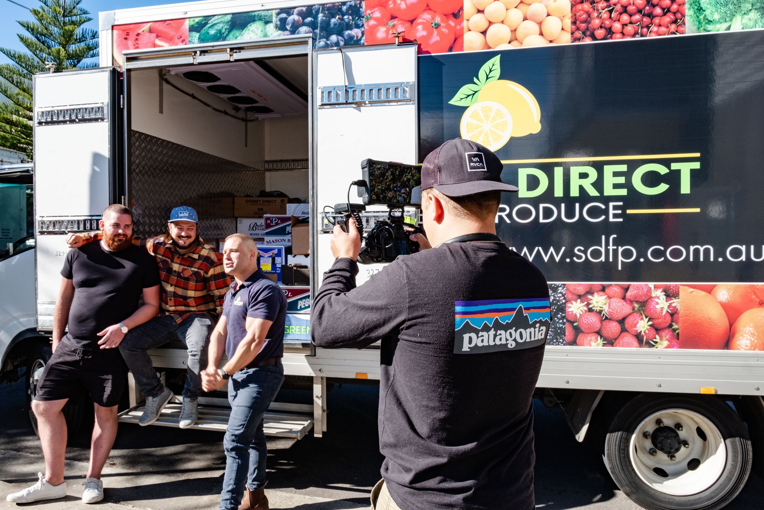 A group of three men posing in front of a produce delivery truck labeled "Direct Produce," while another person films them with a camera. The truck displays images of fruits and vegetables.