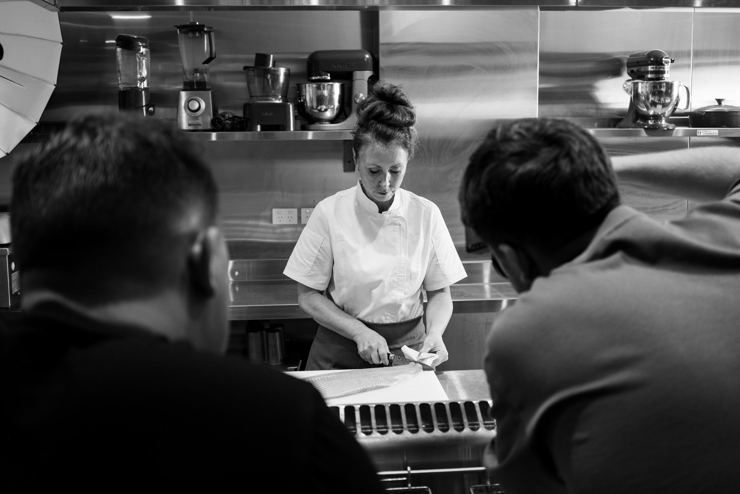 Chef in commercial kitchen preparing fish on cutting board with two people observing, surrounded by kitchen appliances.