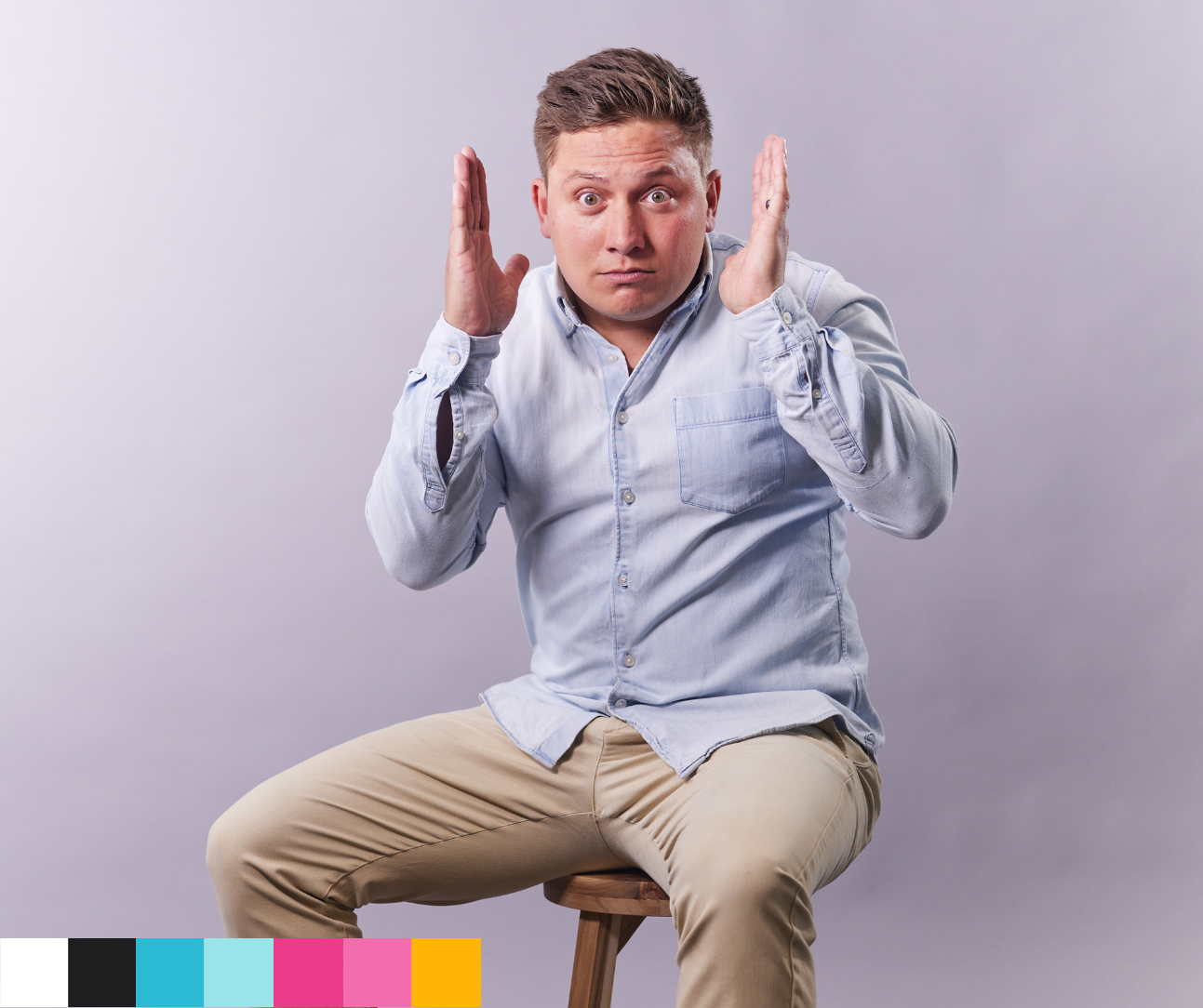 Man with surprised expression, wearing a light blue shirt, sitting on a stool with hands raised, gray background.
