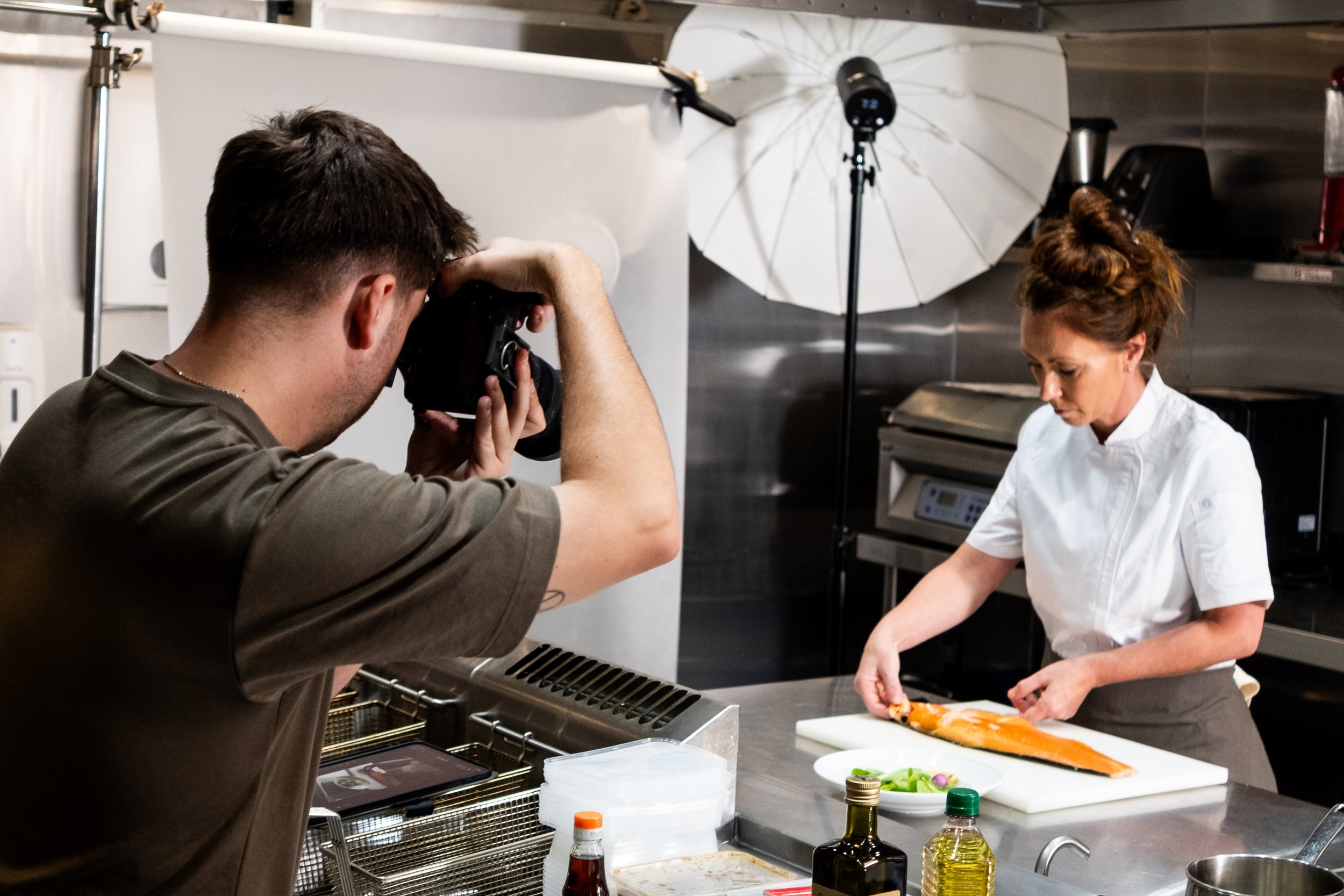 Photographer capturing a chef preparing salmon in a professional kitchen setting.