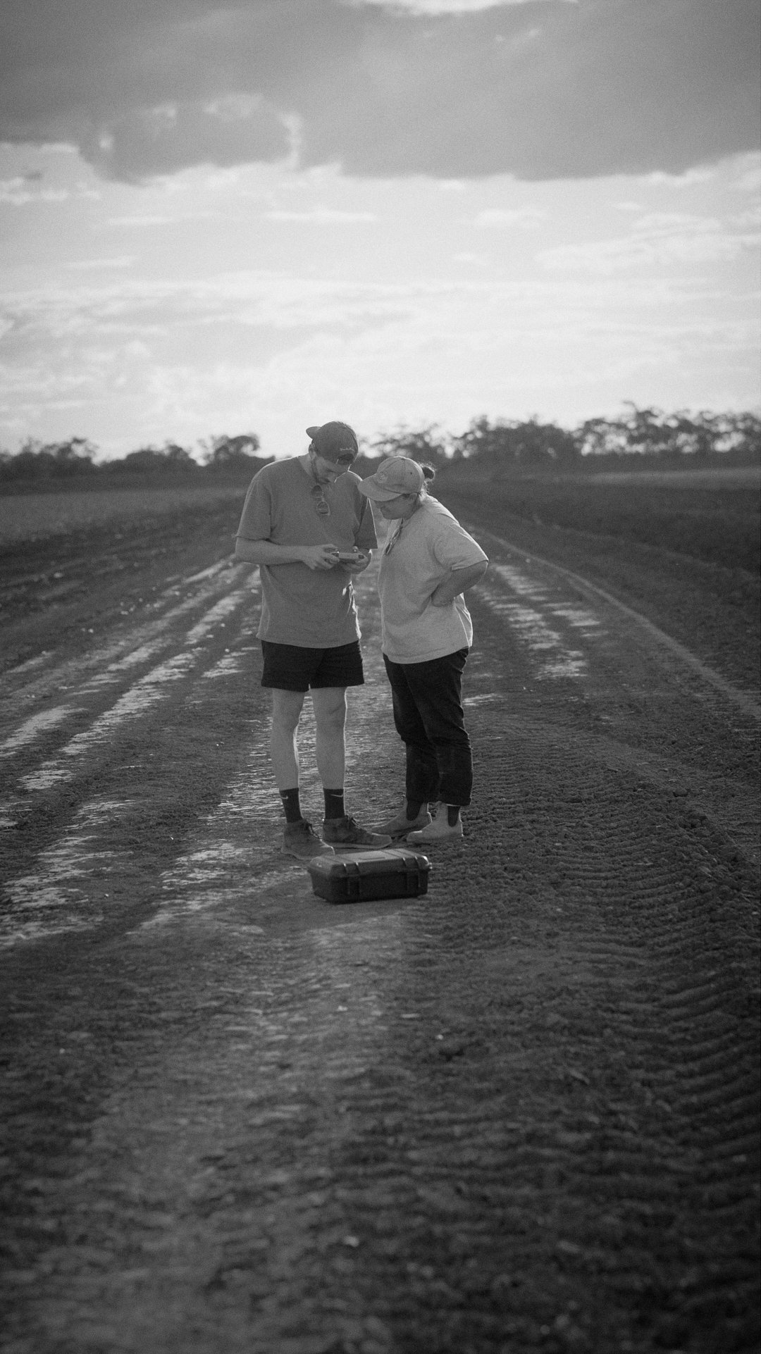 Two people standing on a dirt road, looking at a device, with a closed toolkit on the ground in front of them.