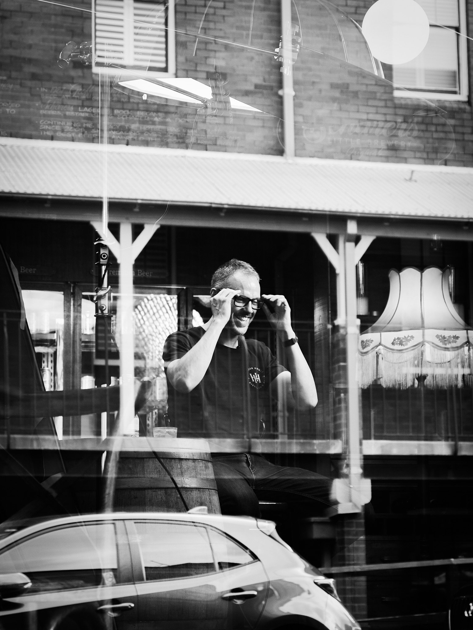 Black and white photo of a man adjusting his glasses, sitting inside a café, viewed through a window with reflections of a car and a building.