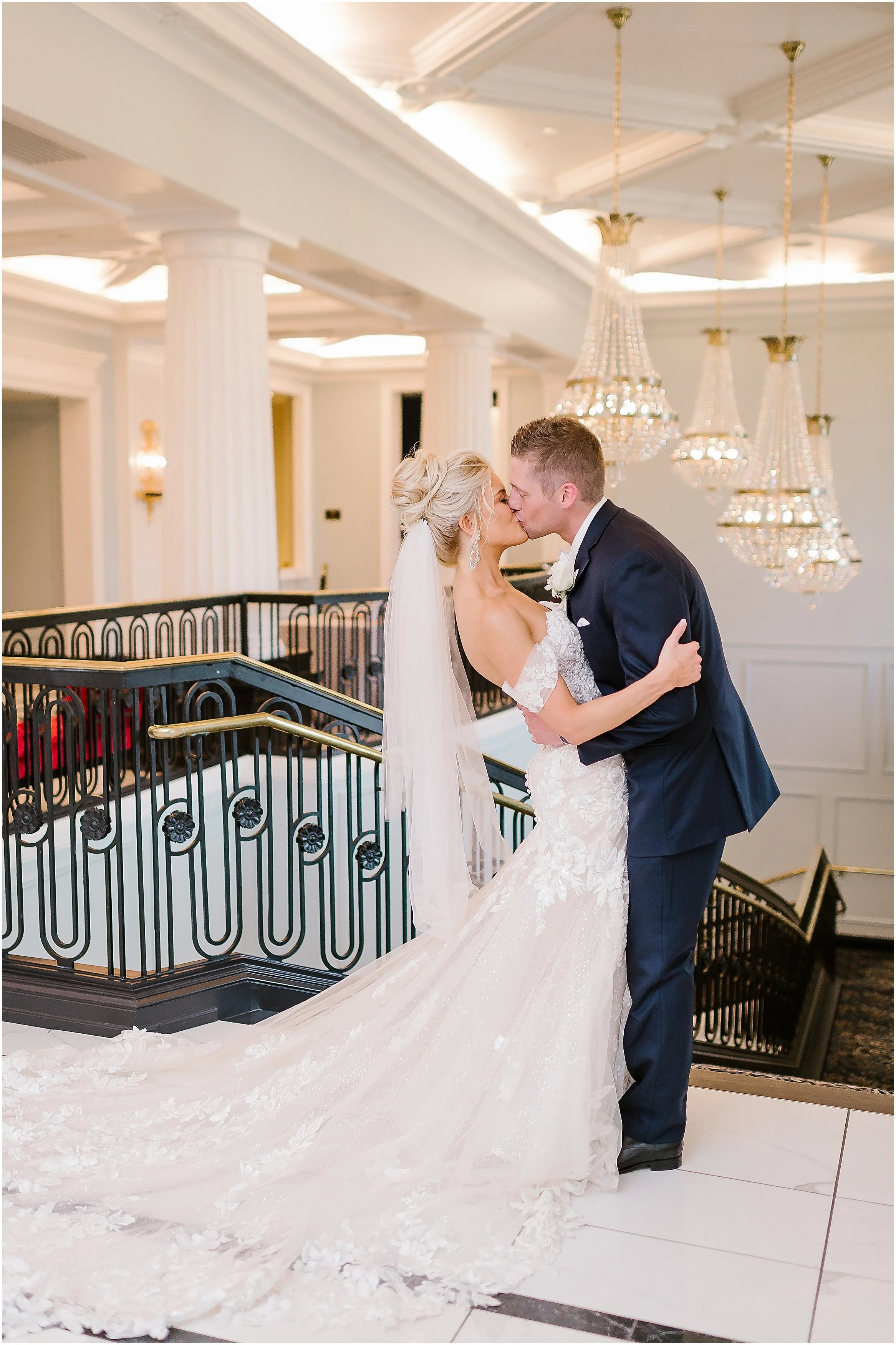 A bride and groom sharing a kiss on a staircase in a grand hall with chandeliers and white columns.