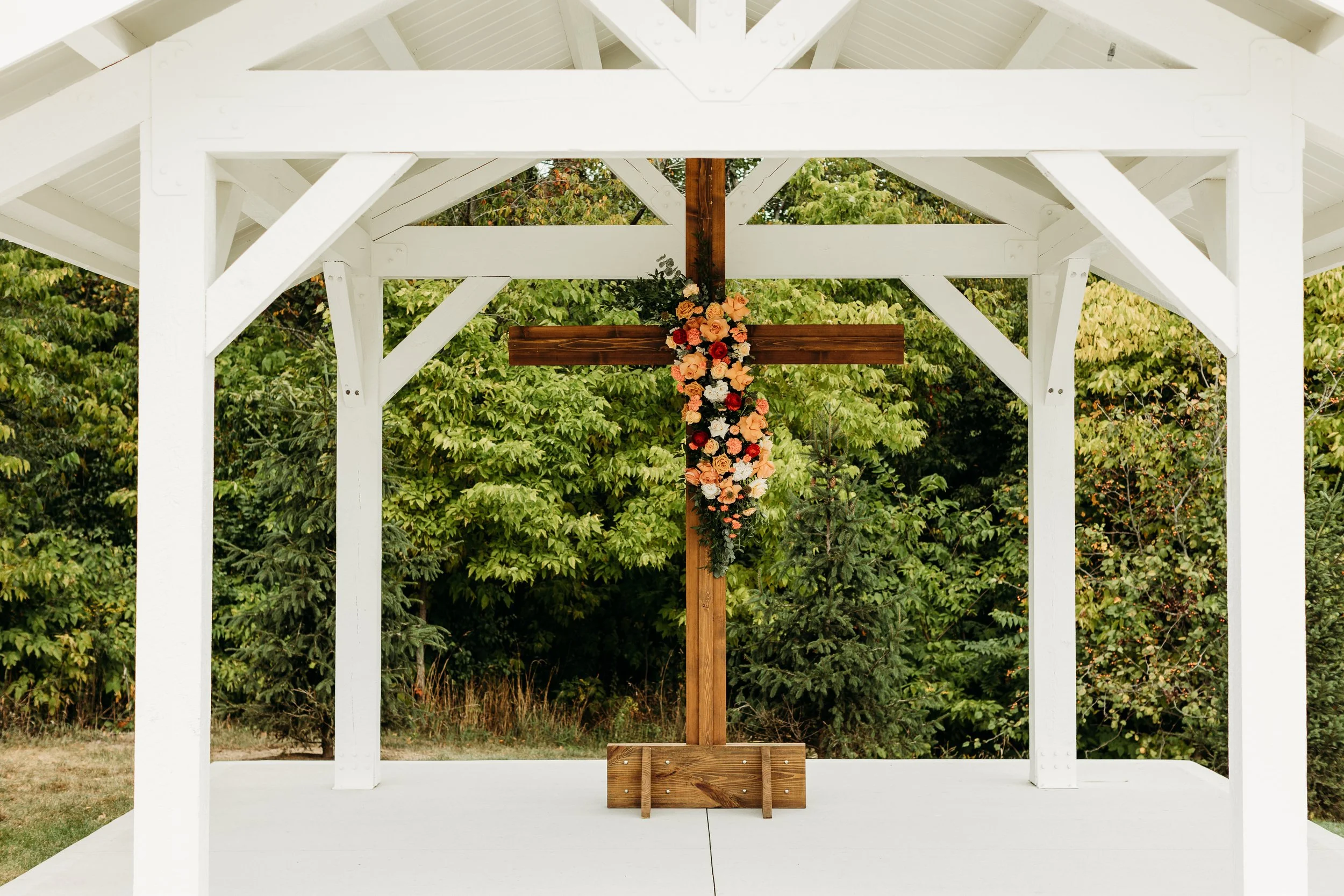 Wooden cross decorated with a floral arrangement under a white pavilion with a natural background of trees.