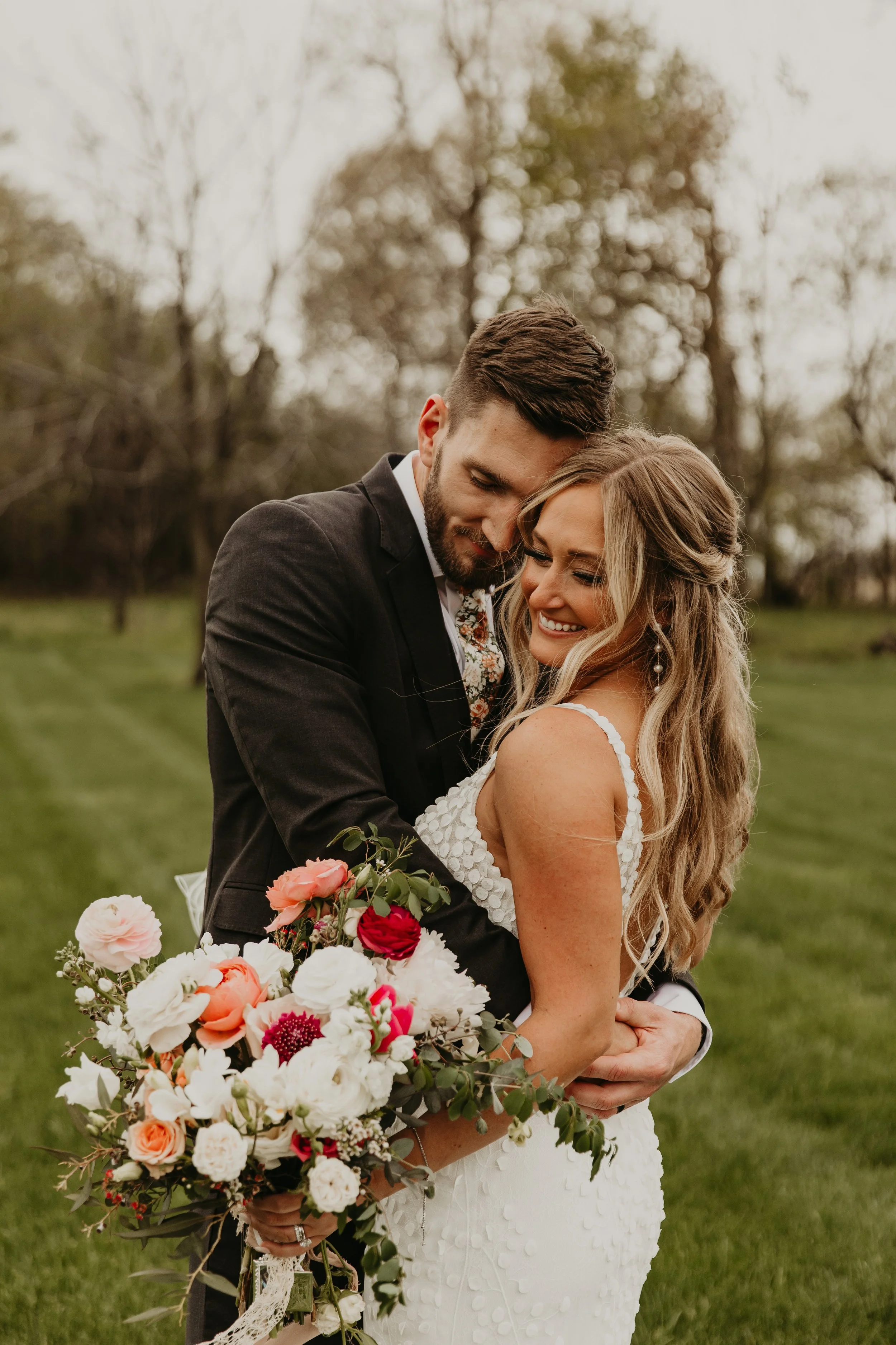 A newlywed couple embraces outdoors, the bride holding a bouquet of colorful flowers, smiling happily.
