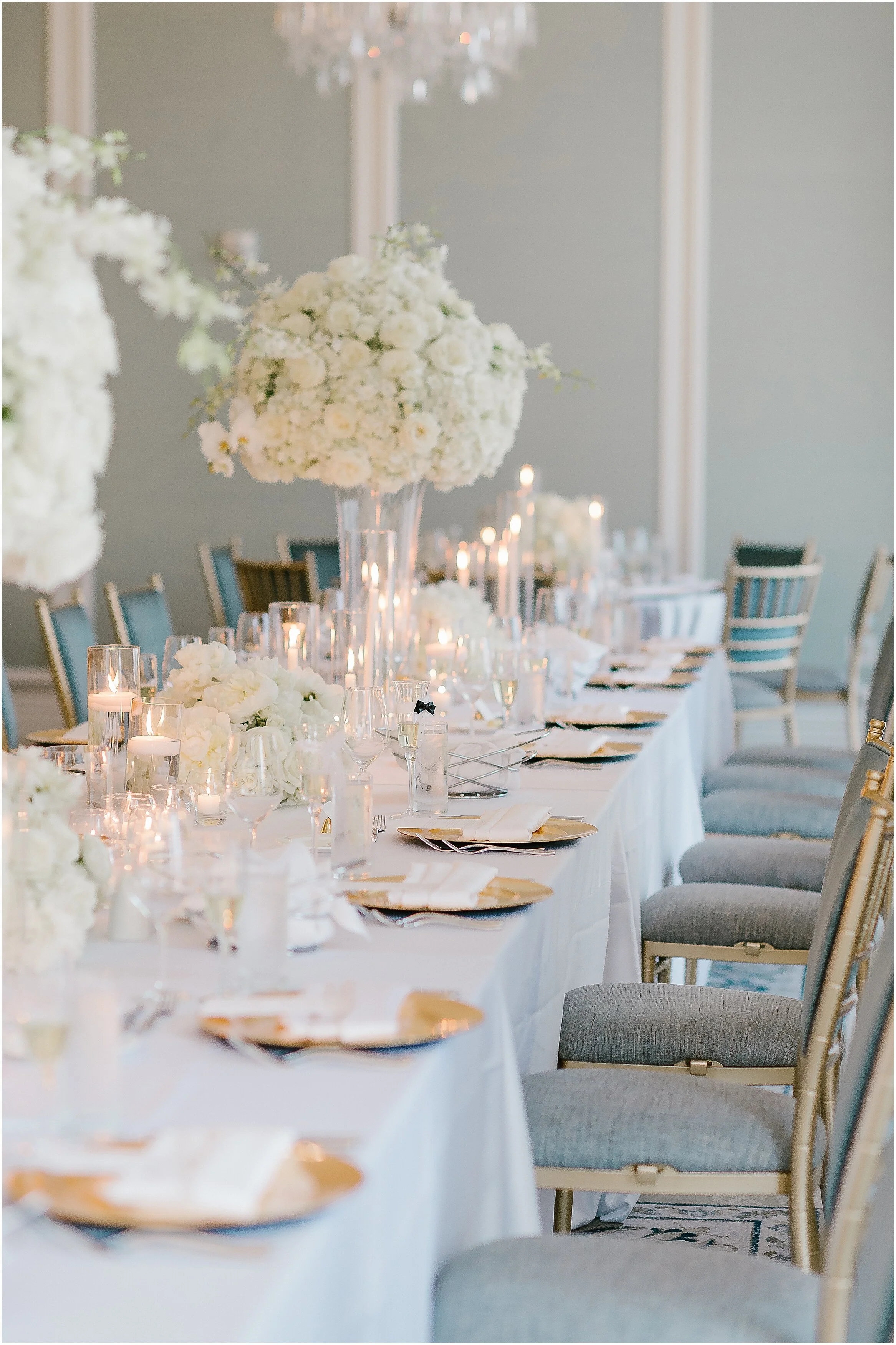 Elegant banquet table decorated with tall glass vases filled with white flower arrangements, surrounded by white tablecloths, gold and white plates, white napkins, and numerous candles in glass holders, set in a sophisticated event hall.