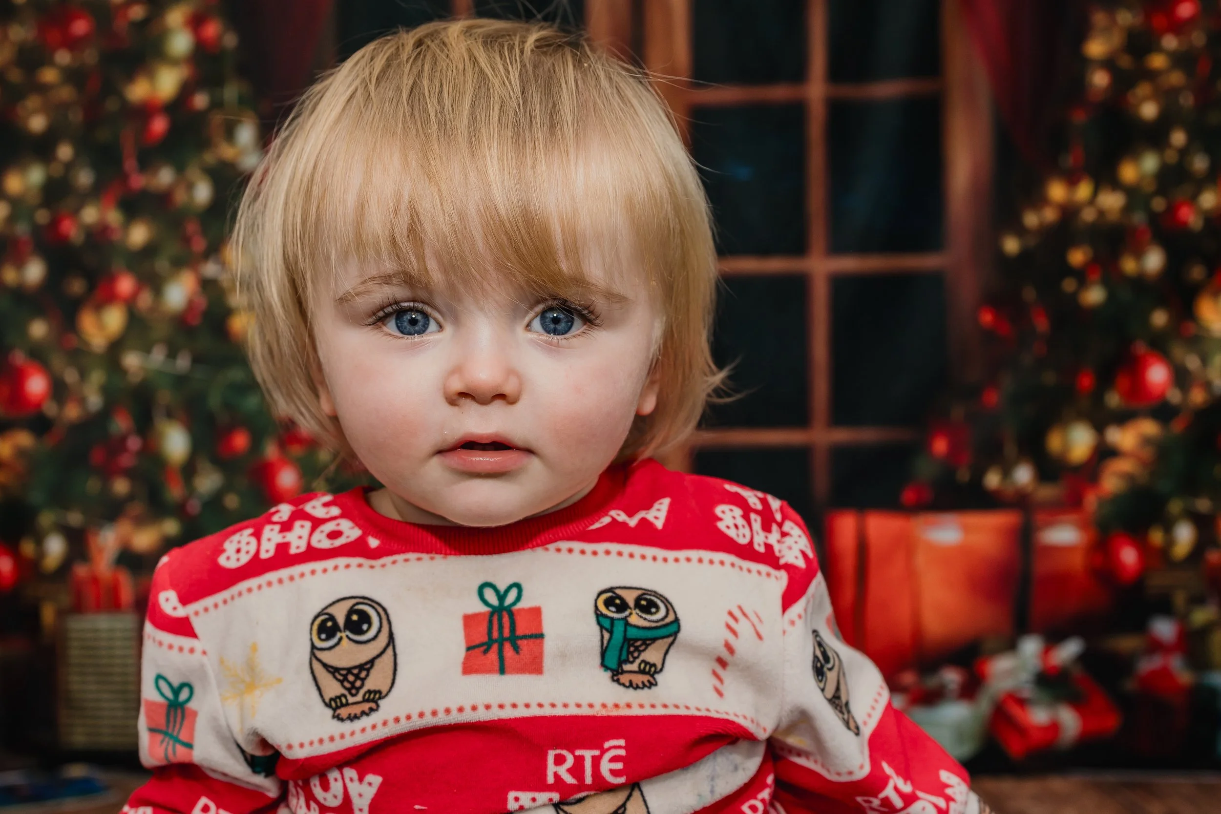 A young child with blonde hair and blue eyes wearing a red and white Christmas-themed shirt, standing in front of a decorated Christmas tree with wrapped presents underneath.