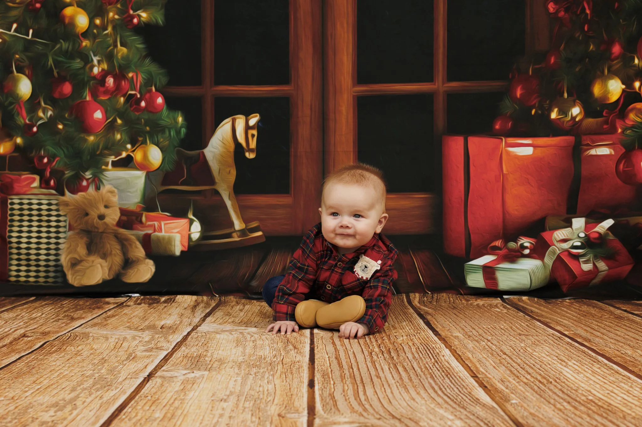 Young child sitting on a wooden floor in front of a Christmas-themed backdrop with presents, Christmas tree, rocking horse, teddy bear, and holiday decorations. Captured in Dublin by family photographer