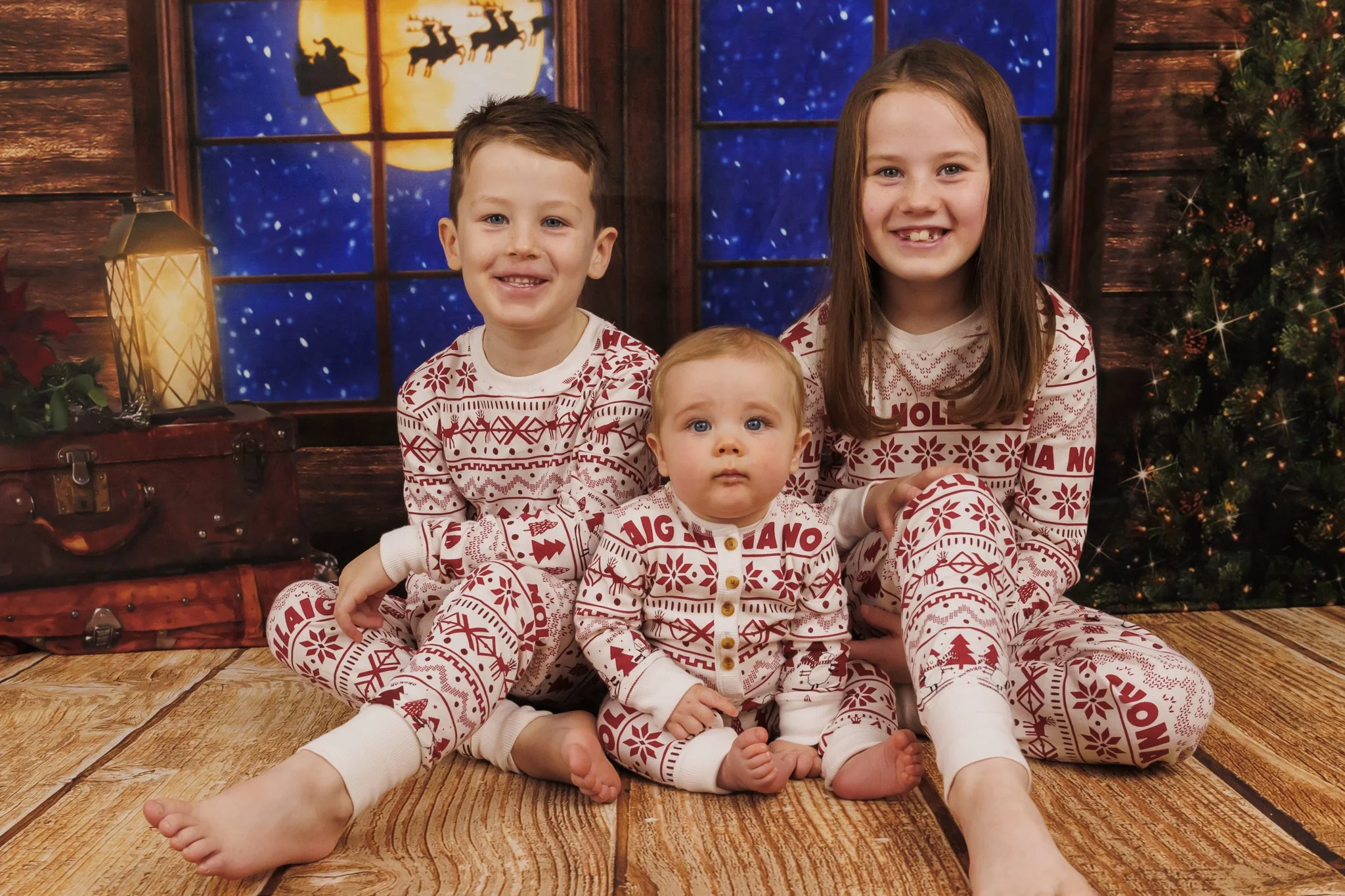 Three children sitting on a wooden floor in Christmas pajamas with a Christmas tree and a window showing a snowy night and Santa's sleigh in the background. Dublin family photographer