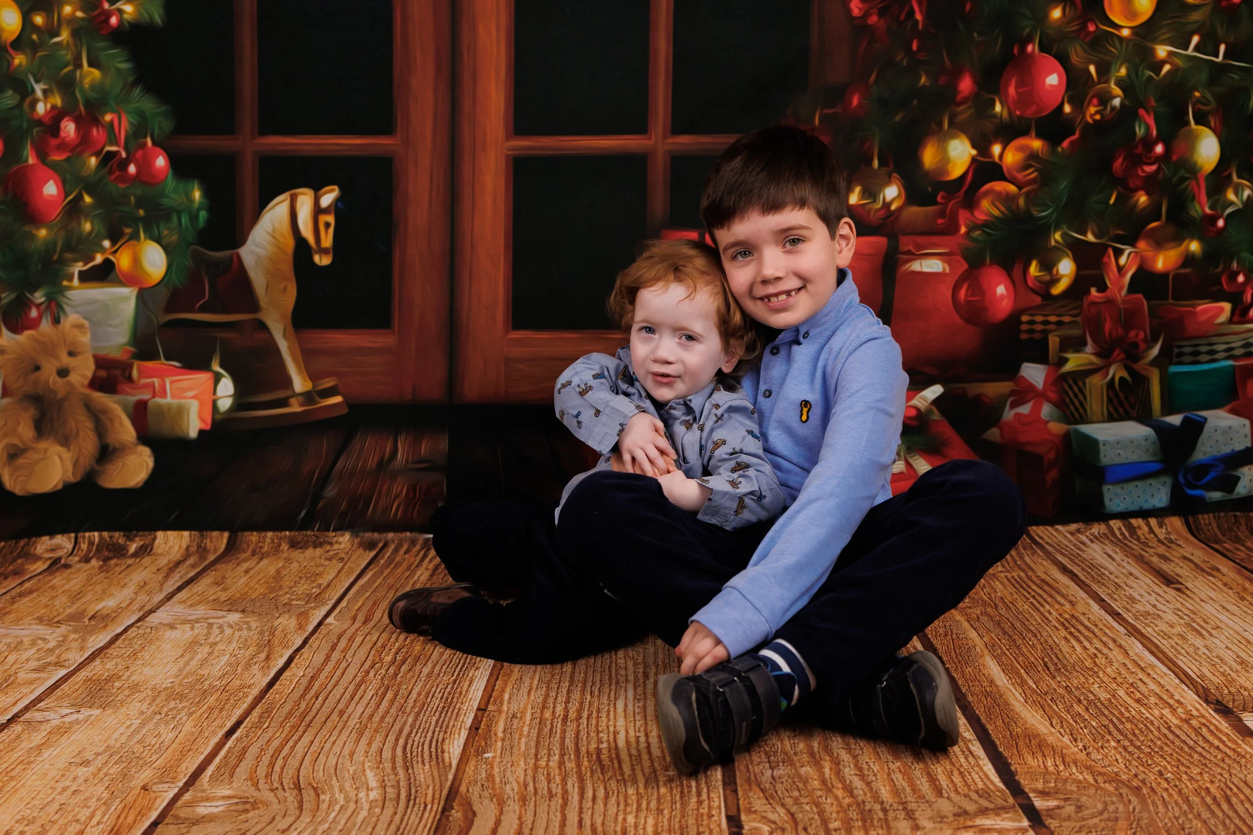 Two children sitting on a wooden floor in front of a Christmas-themed backdrop, with Christmas trees, ornaments, and presents.