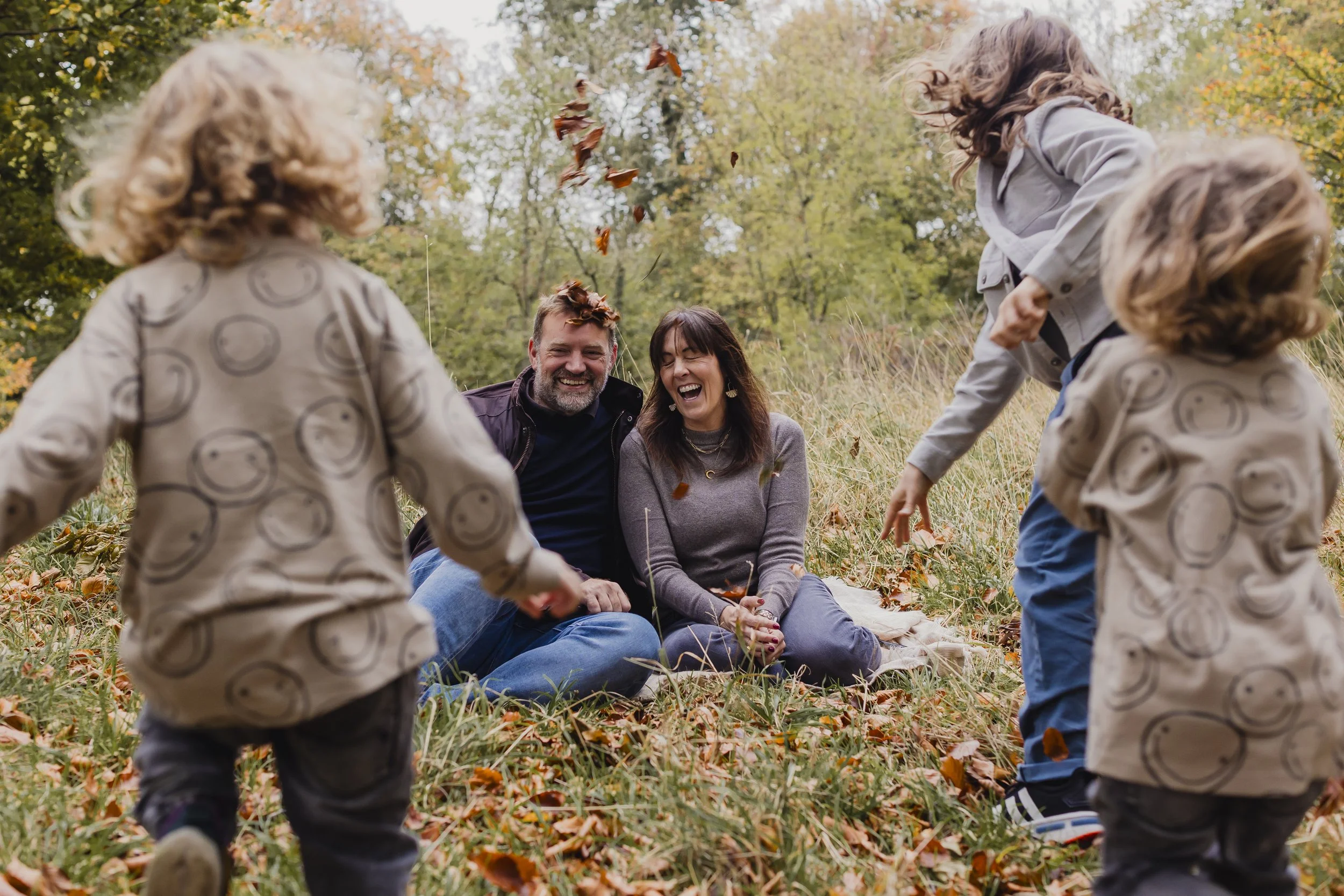 A family enjoying a playful moment outdoors on a fall day, with leaf-throwing among children and parents sitting on a blanket amidst autumn foliage.