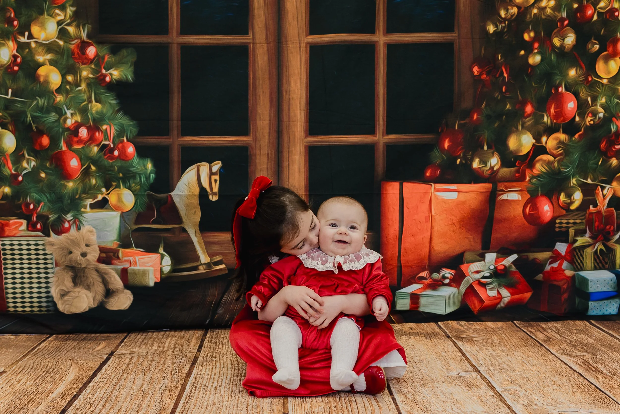 A young girl dressed in a red dress with a red bow kissing a smiling baby, who is also dressed in a red outfit with a white lace collar, sitting on a wooden floor in front of a Christmas-themed backdrop featuring decorated Christmas trees, wrapped gi