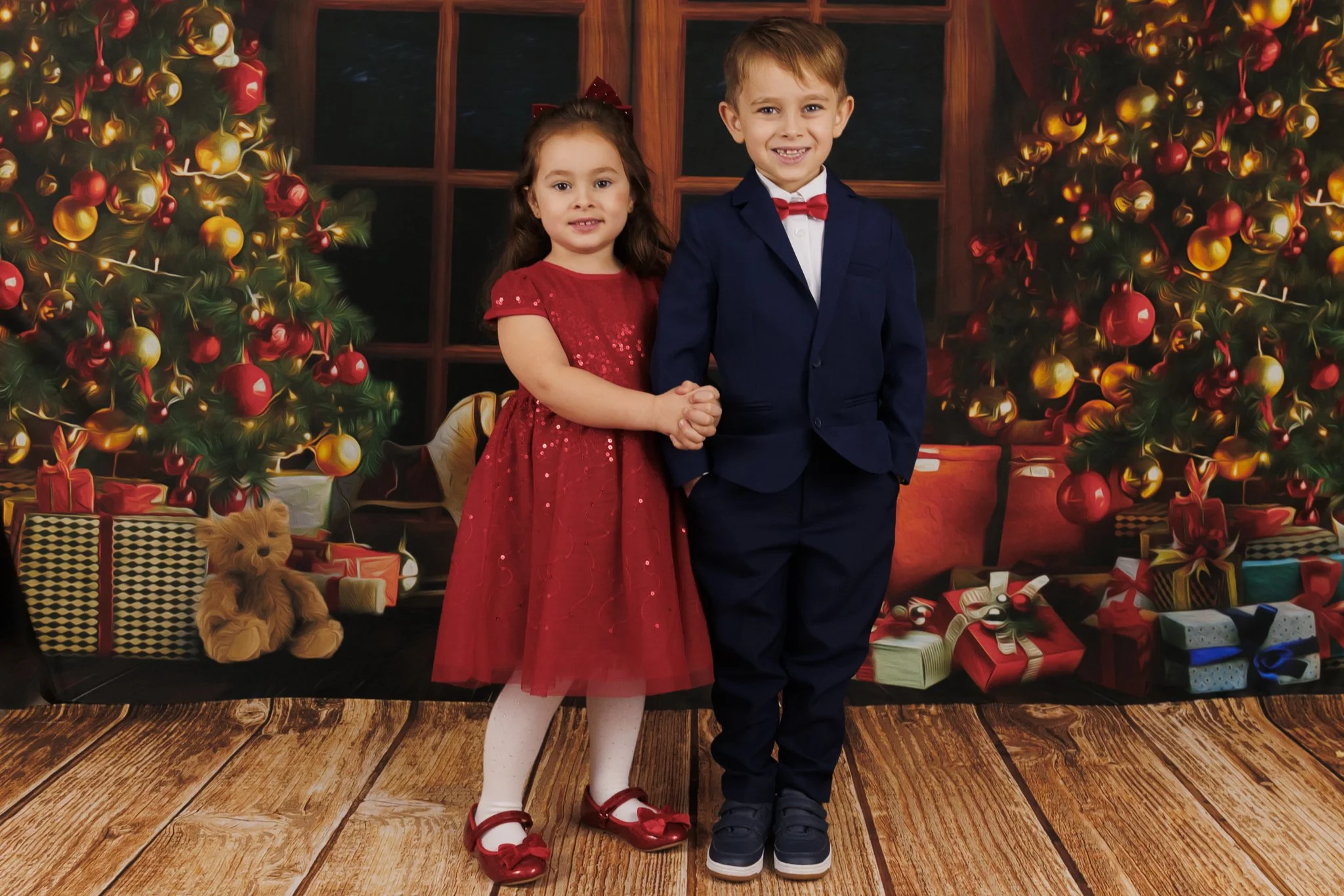 Two children, a girl in a red dress and a boy in a navy suit, pose in front of a Christmas-themed backdrop with decorated Christmas trees and presents.