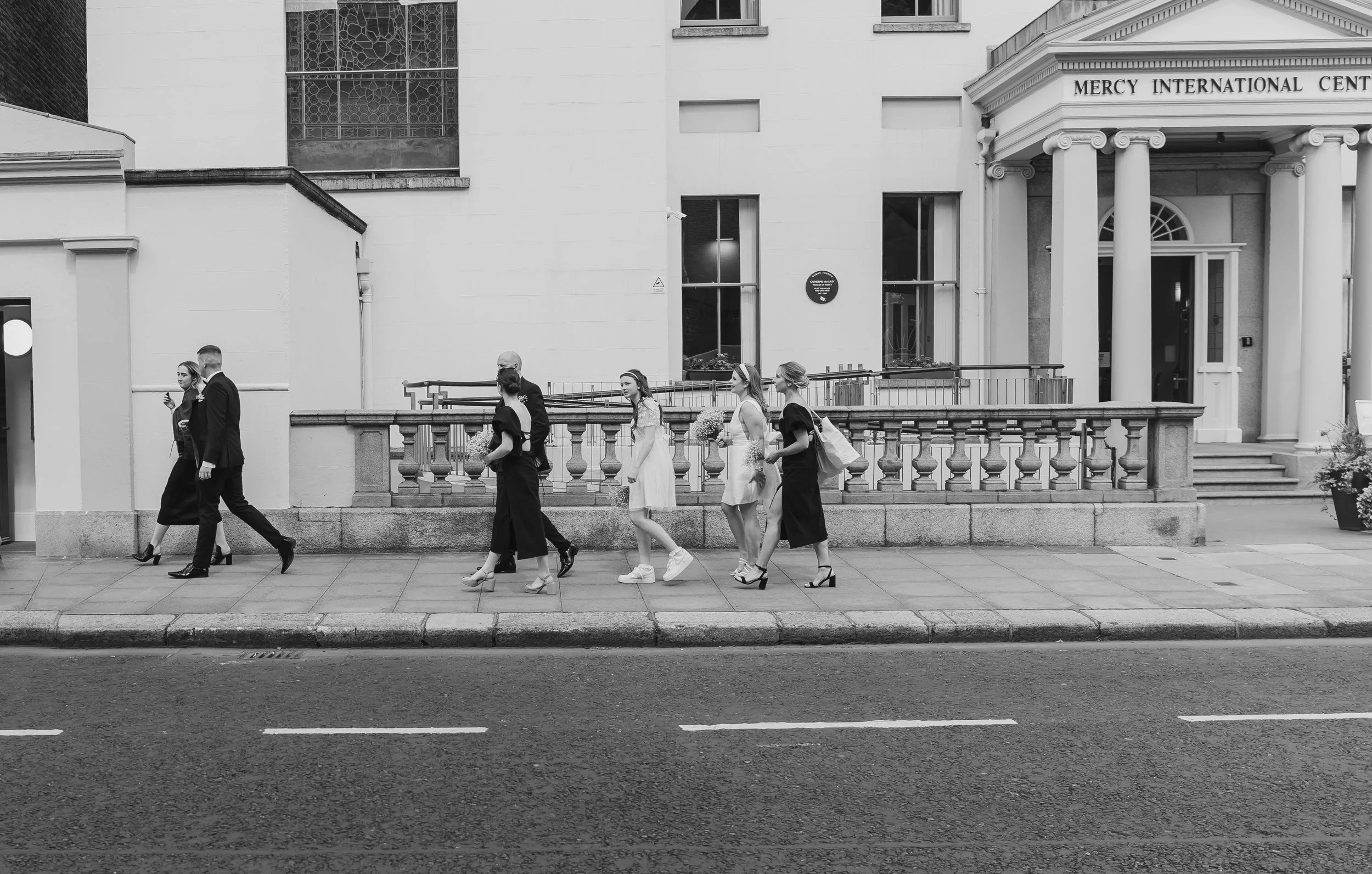 Black and white photo of seven people walking on a sidewalk in front of a building with the sign Mercy International Center. Four women in dresses and heels, and three men dressed in suits. Some women are carrying flowers and bags while one woman is looking at her phone.