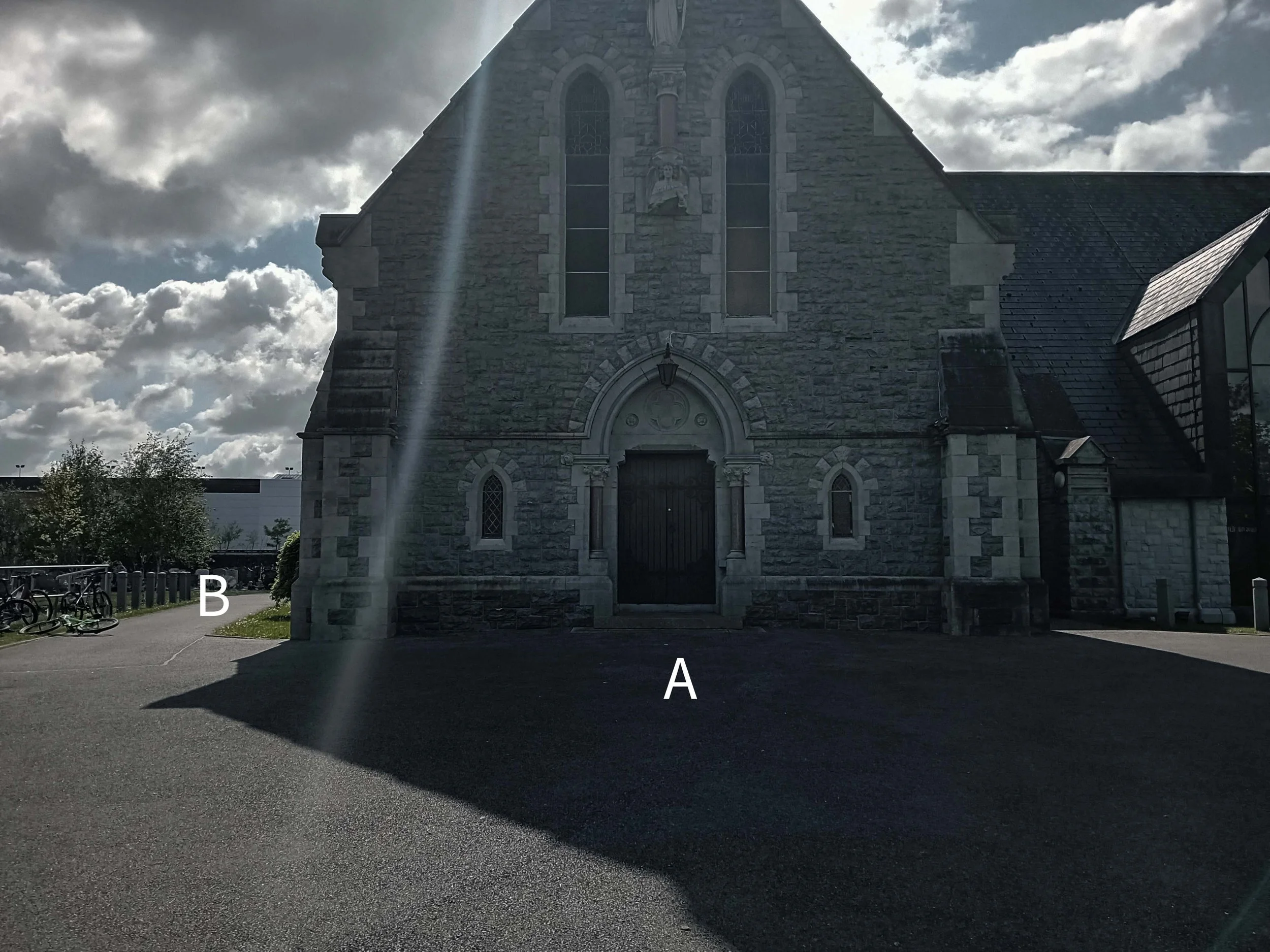 A stone church with pointed stained glass windows and a dark door, with a bright light shaft hitting the front. Bicycles are parked on the left, and the church's shadow is cast on the ground. A shadowed parking lot is in the foreground.