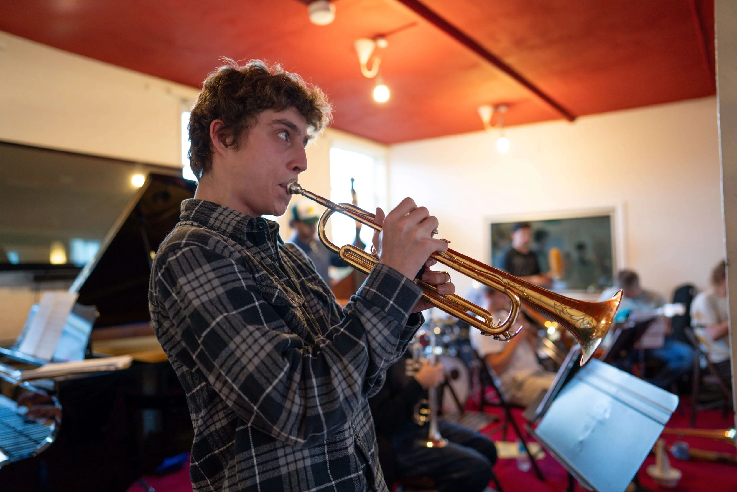 Trumpeter rehearsing with peers; piano and music stands behind.