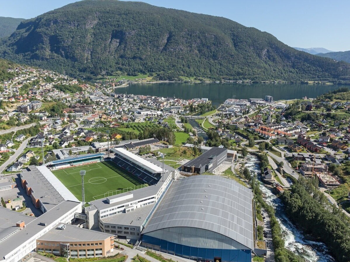 Luftfoto av en by med et fotballstadion i forgrunnen, omgitt av fjell og en innsjø i bakgrunnen. Det er mange bygninger og boliger spredt rundt i området.