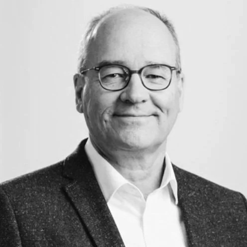 A black-and-white portrait of a middle-aged man with glasses, wearing a suit jacket and white shirt, smiling.