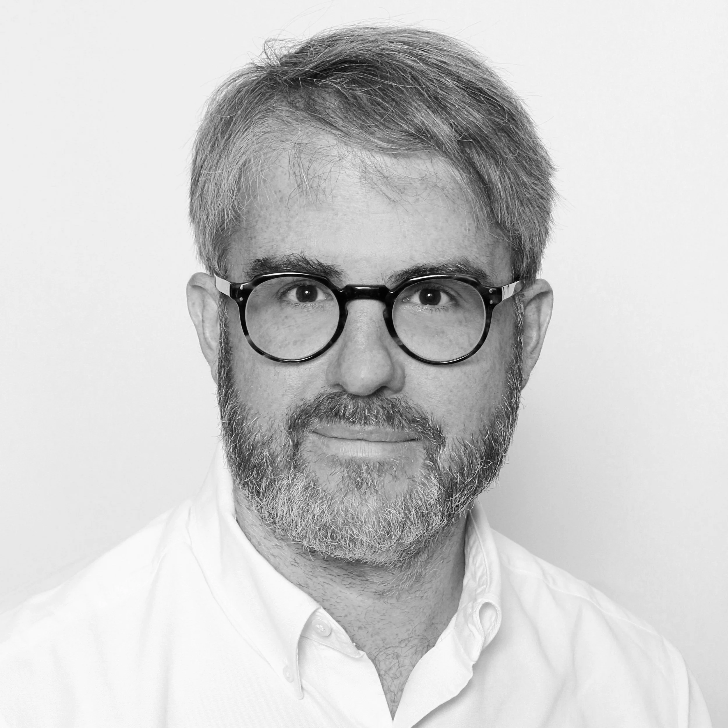 Black and white headshot of a man with glasses, a beard, and short, slightly tousled hair, wearing a white collared shirt.