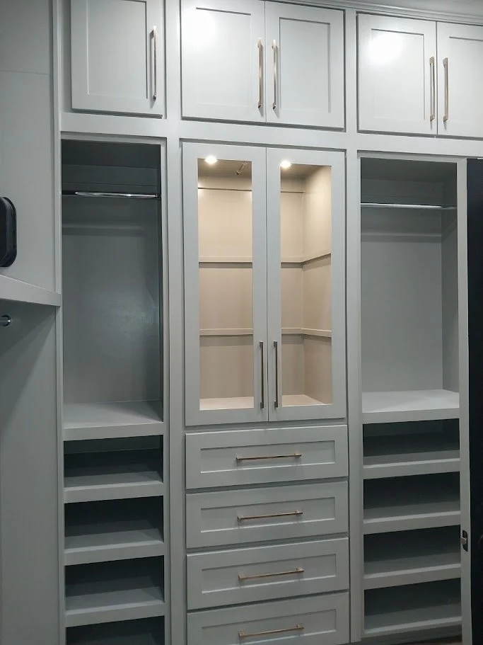 Custom walk-in closet built by Greywolf Cabinets and More, featuring light gray built-in cabinetry, hanging rods, glass-front display shelves, and integrated drawers with sleek hardware.