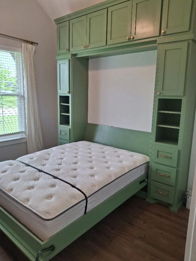 Custom sage green Murphy bed folded down in the open position, built by Greywolf Cabinets and More with matching surrounding storage cabinetry, drawers, and open shelving.