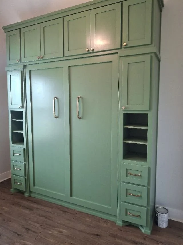Custom sage green Murphy bed closed in a wall unit built by Greywolf Cabinets and More, featuring upper cabinets, side shelving towers, and drawers with brass hardware.