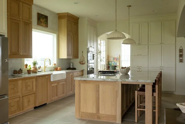 A beautifully lit, custom light-wood kitchen showcasing floor-to-ceiling cabinetry by Greywolf Cabinets and More, a large central window above a farmhouse sink with brass faucet. a light stone countertop, and integrated stainless steel double ovens.