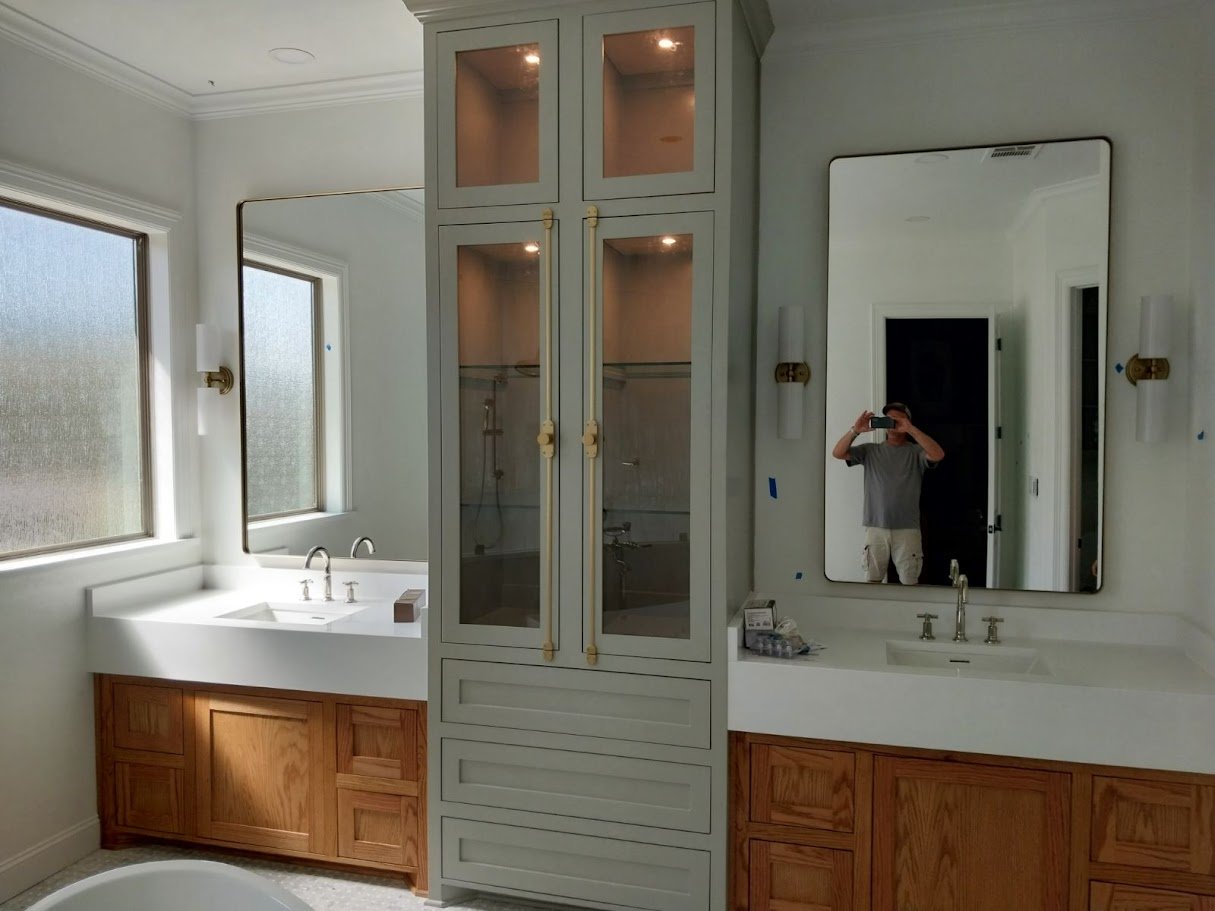 Custom bathroom vanity and matching floor-to-ceiling linen tower built by Greywolf Cabinets and More, featuring dual sinks, natural wood lower cabinets, and warm gray upper cabinetry.