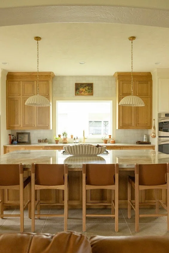 Custom light wood kitchen cabinets and a matching large island with leather-upholstered barstools, built-in upper cabinetry flanking a large bright window, and modern brass pendant lighting.