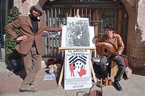 Due uomini sul marciapiede con un cartellone pubblicitario per uno spettacolo teatrale, uno con chitarra e l'altro con un berretto, davanti a un cancello in ferro e a un poster con l'immagine di due persone sedute.