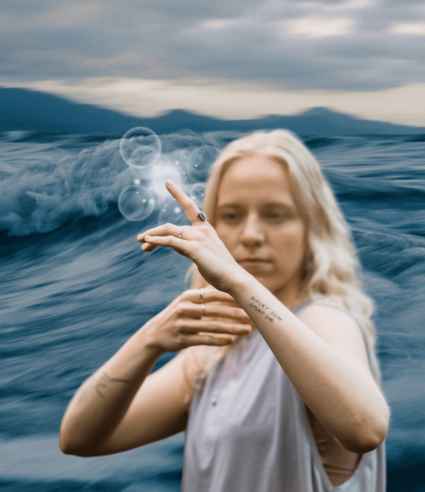 Eine blonde Frau in einem weißen Top mit Tattoos auf den Armen und mehreren Ringen auf den Fingern steht vor einer Meereskulisse und berührt eine digitale, leuchtende Blase, die sich in der Luft befindet.
