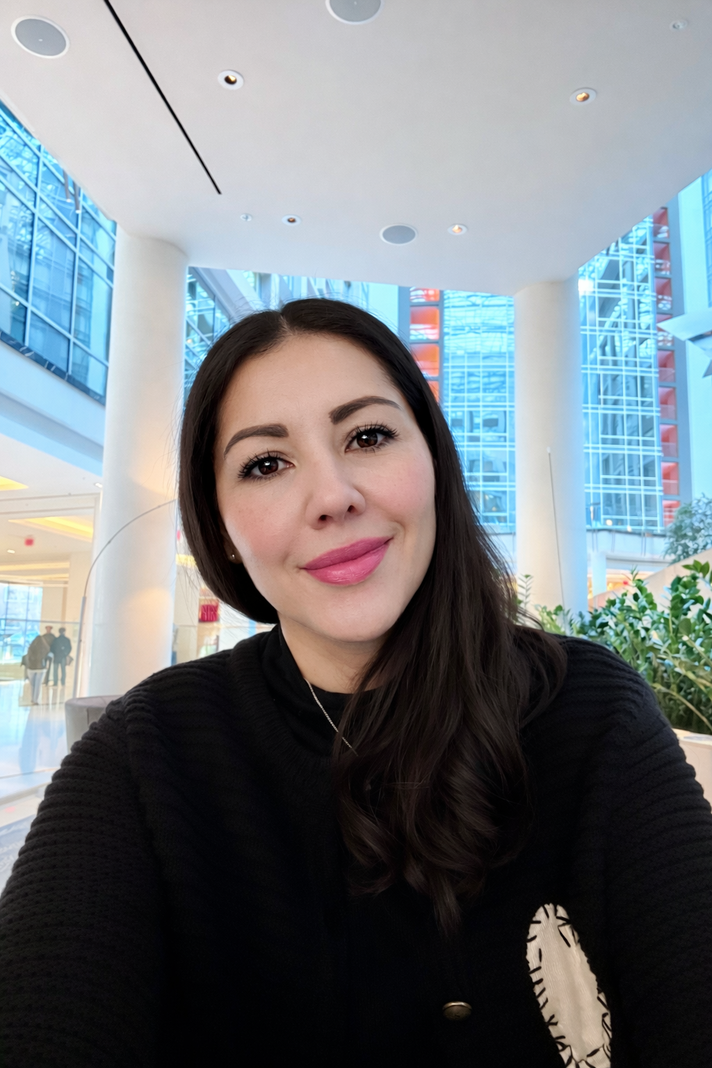 A woman with long dark hair, wearing a black sweater, smiling in an indoor space with large glass windows and modern architecture.