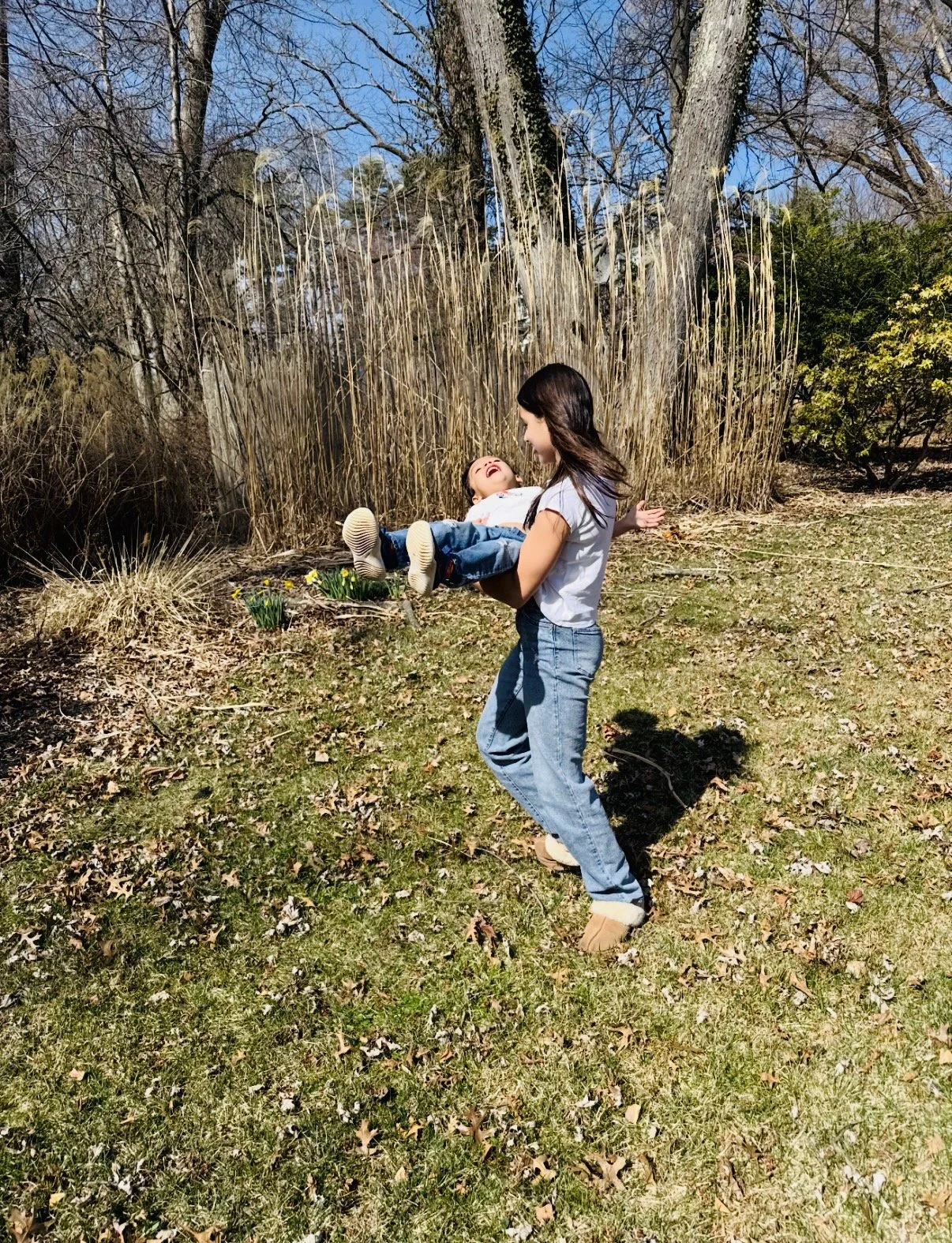 A young girl in blue jeans and a white T-shirt holding a laughing child in a garden with tall dried grass and leafless trees.