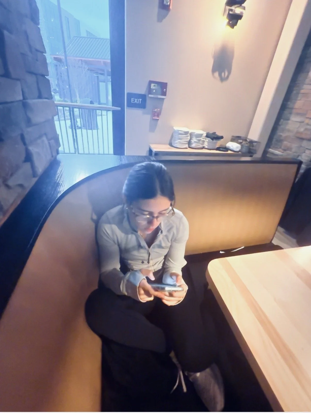 A woman sitting on a bench in a café, looking at her smartphone with a focused expression. The café has a wooden table and brick walls, with a view of an outdoor balcony through a glass door.