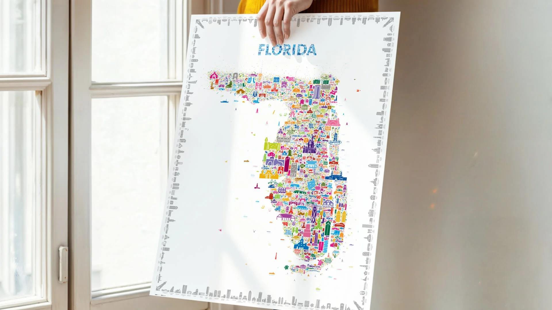 Person holding the Iconic Florida Poster near a window, showing the full colorful illustrated map of Florida with hand-drawn icons by Rafael Esquer for Alfalfa Studio.