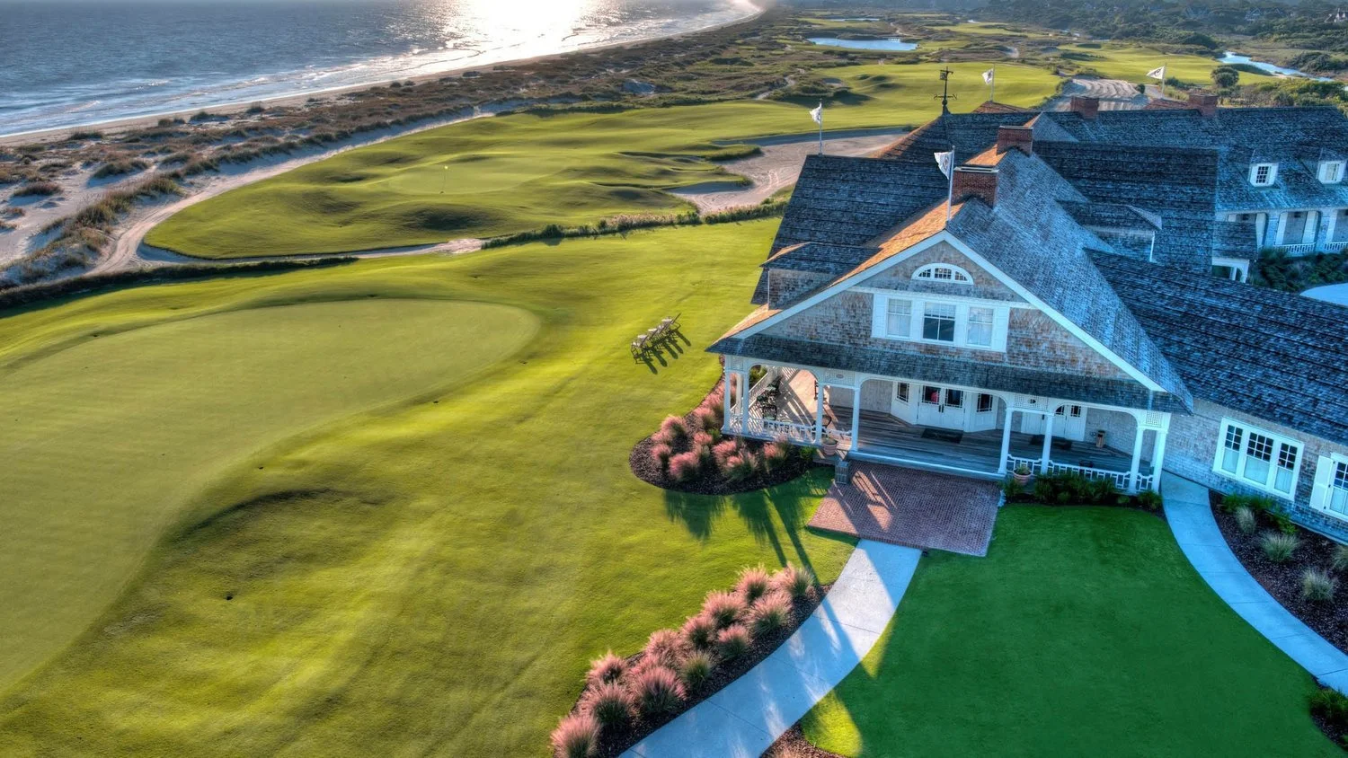 A large house with a porch, situated next to a golf course with green fairways and sand traps, near the coastline.