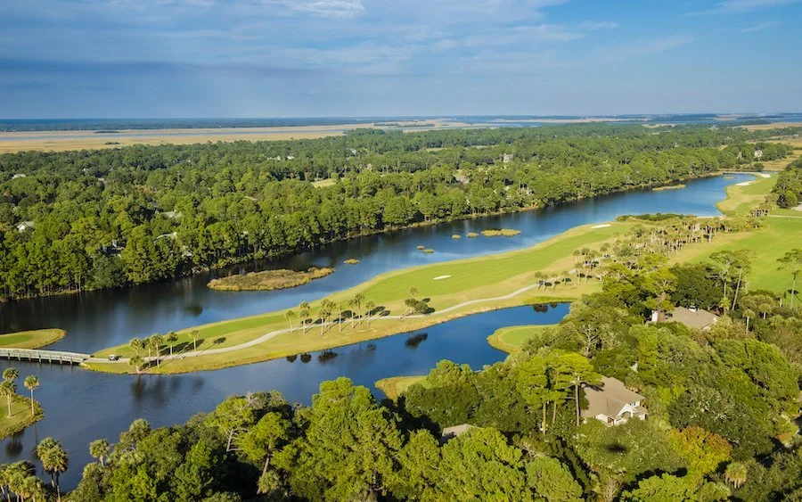 Aerial view of a golf course with green fairways, trees, water hazards, and a bridge, surrounded by forested areas and distant fields.